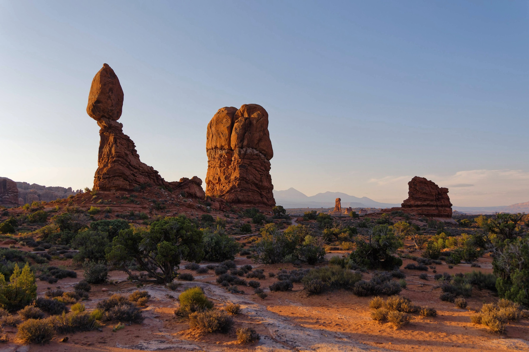 A desert landscape featuring several large rock formations. The leftmost one resembles a giant egg-shaped rock balanced on a pedestal. 