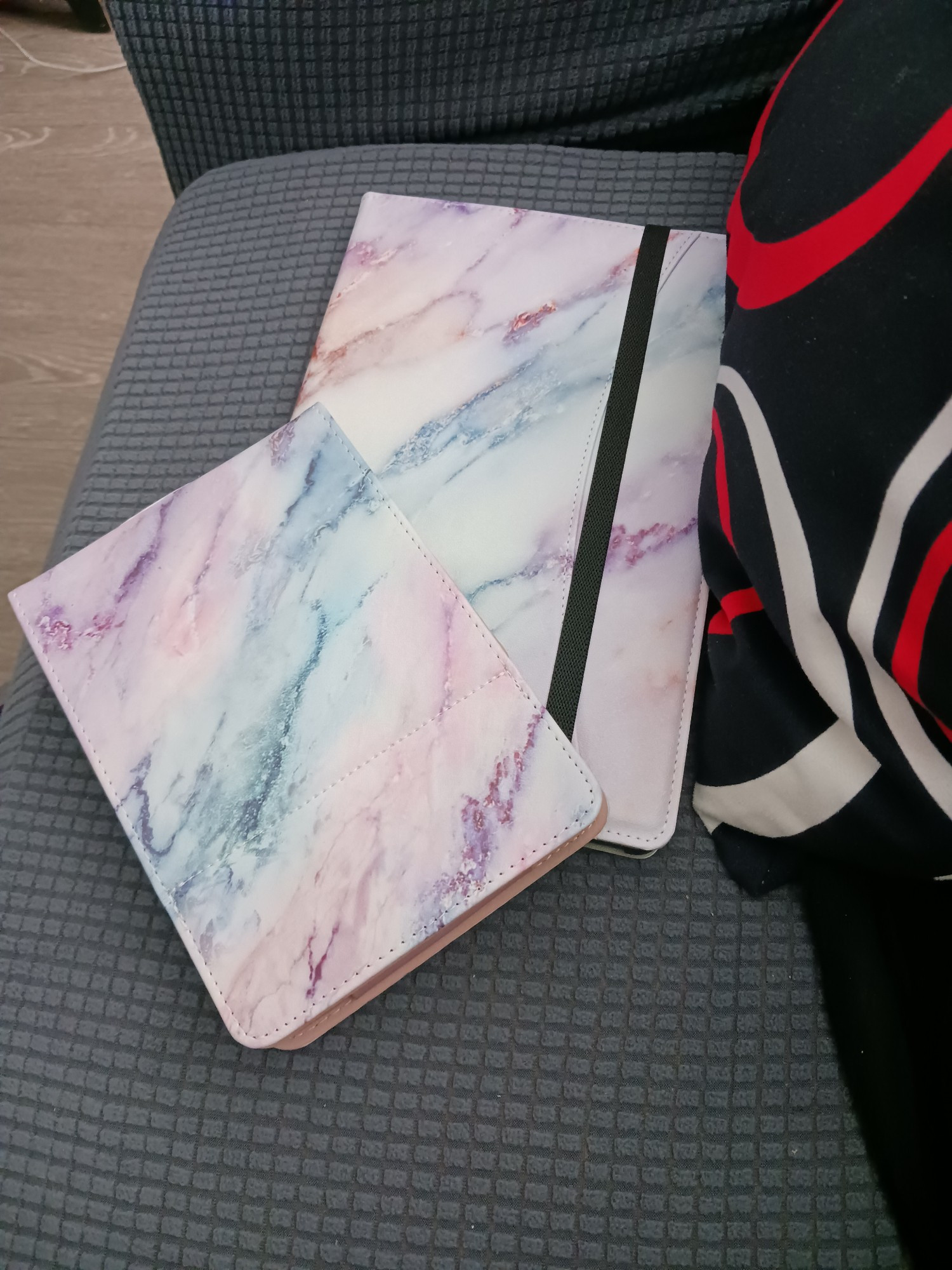 Grey background. 2 rectangular tablet covers, both marbled pink/grewn/purple, next to a black/red/white pillow.