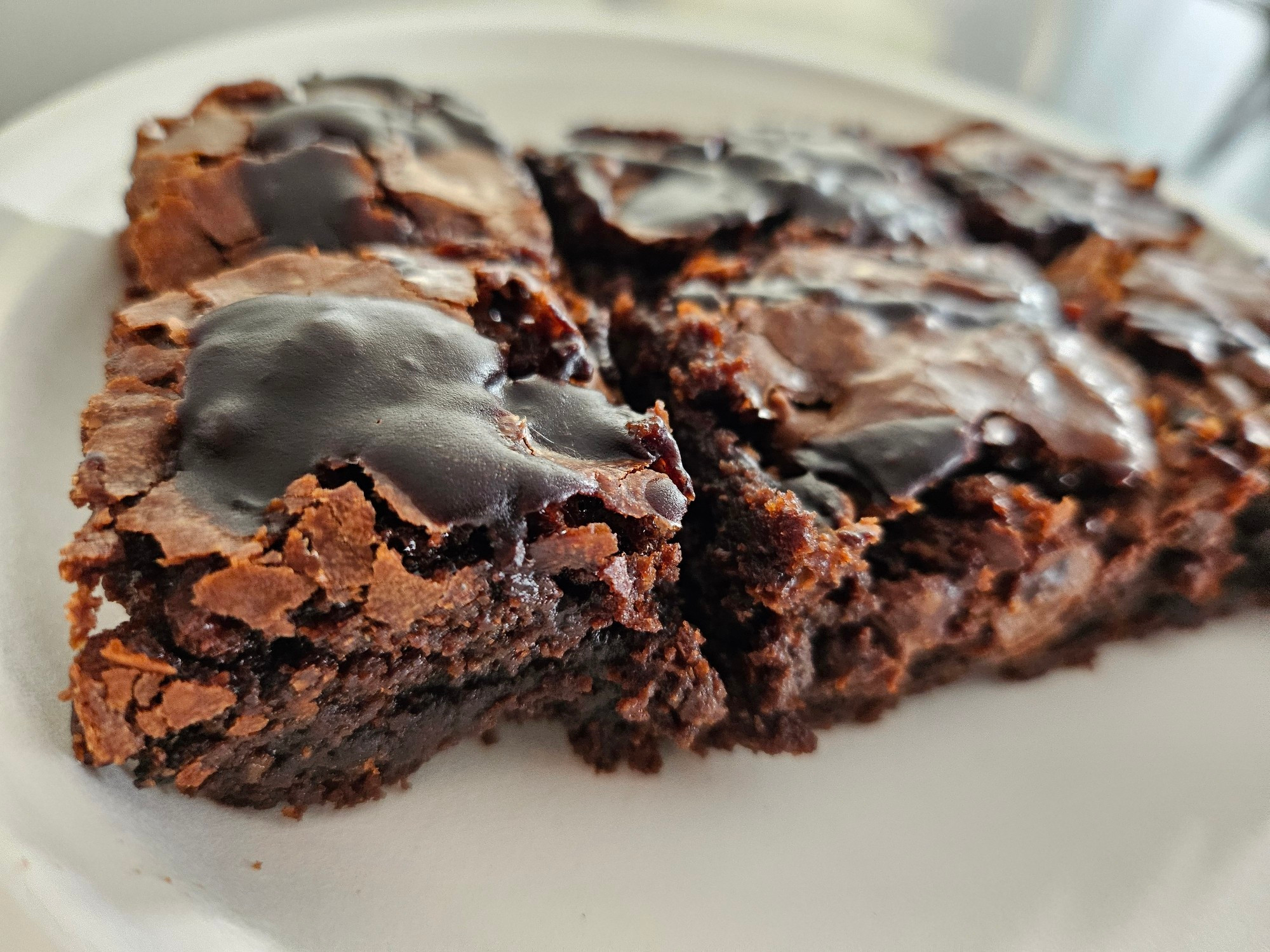 A close up photograph of chocolatey brownies with a chocolate ganache splattered over the top. The top layer is flakey, and the bottom layer is oh so fudgy.