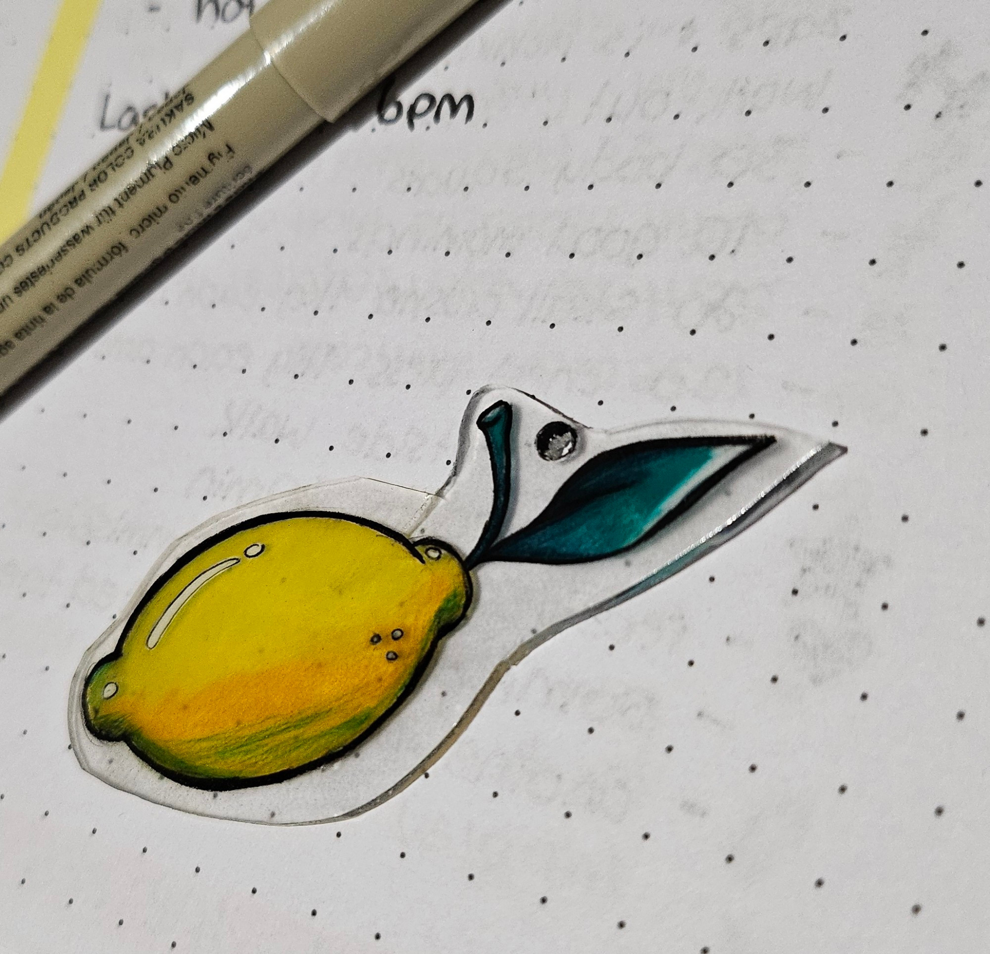 A photo of a hand drawn lemon shrinky dink. It sits on a page of dotted paper with a pen in the background.