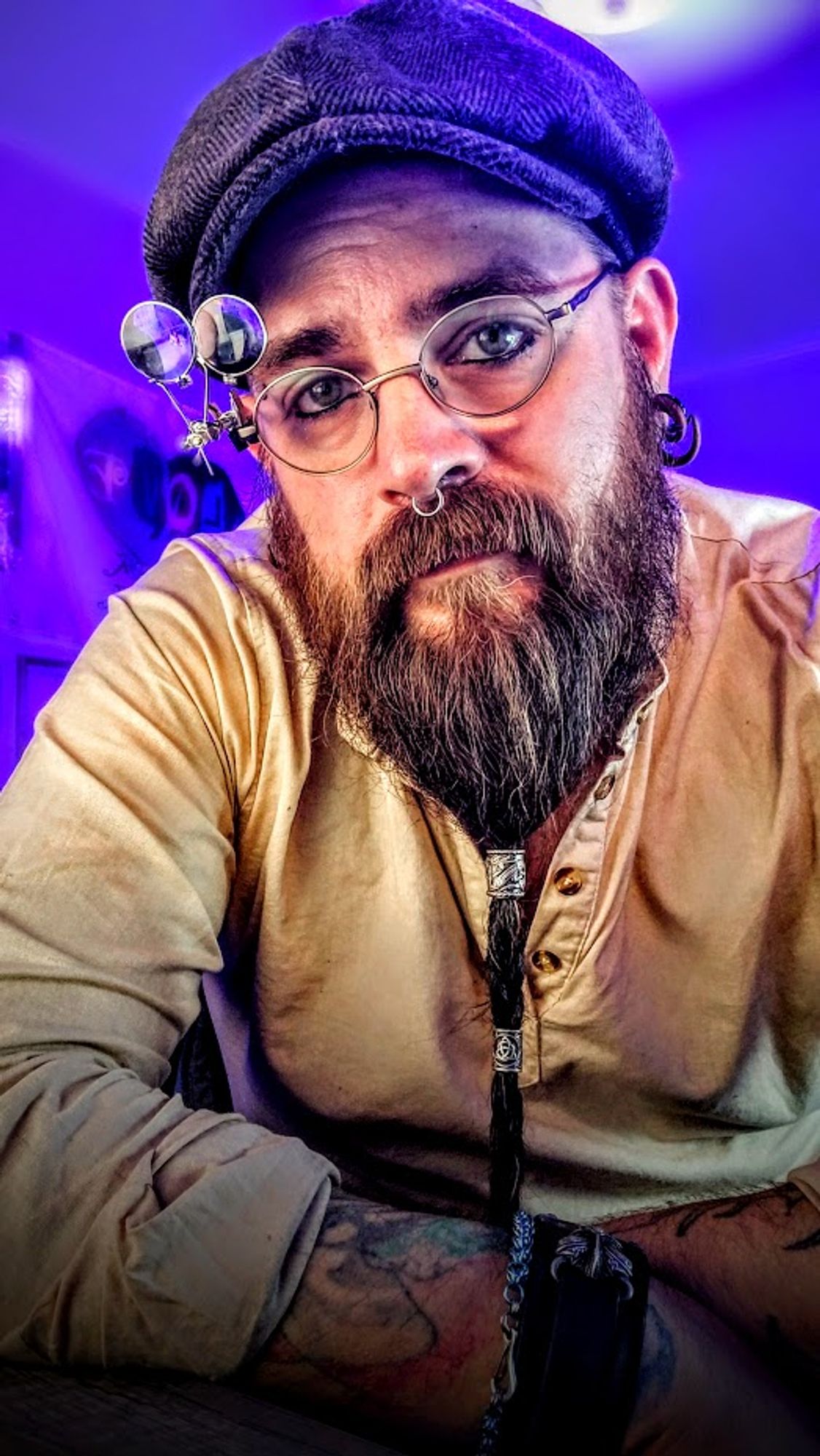 Man with a long braided beard wearing round spectacles with two magnifying lenses attached. He has on a flat cap and tan tunic