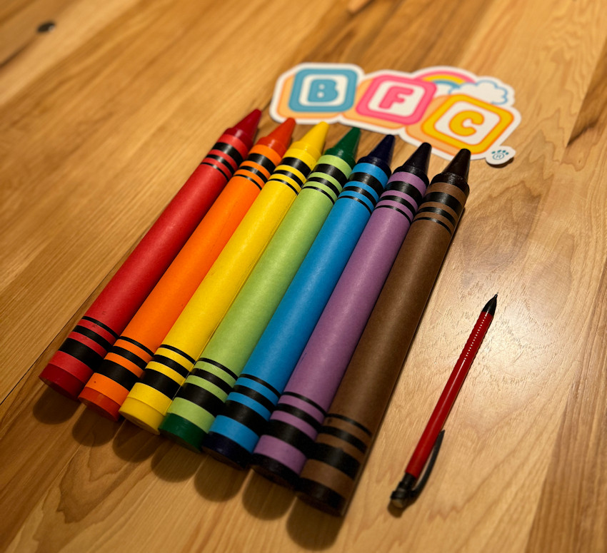 Red, Orange, Yellow, Green, Blue, Purple, and Brown Jumbo Sized Crayons laying on a table next to a Pencil, and a B F C Sticker for scale. The Crayons are about 10 times larger.