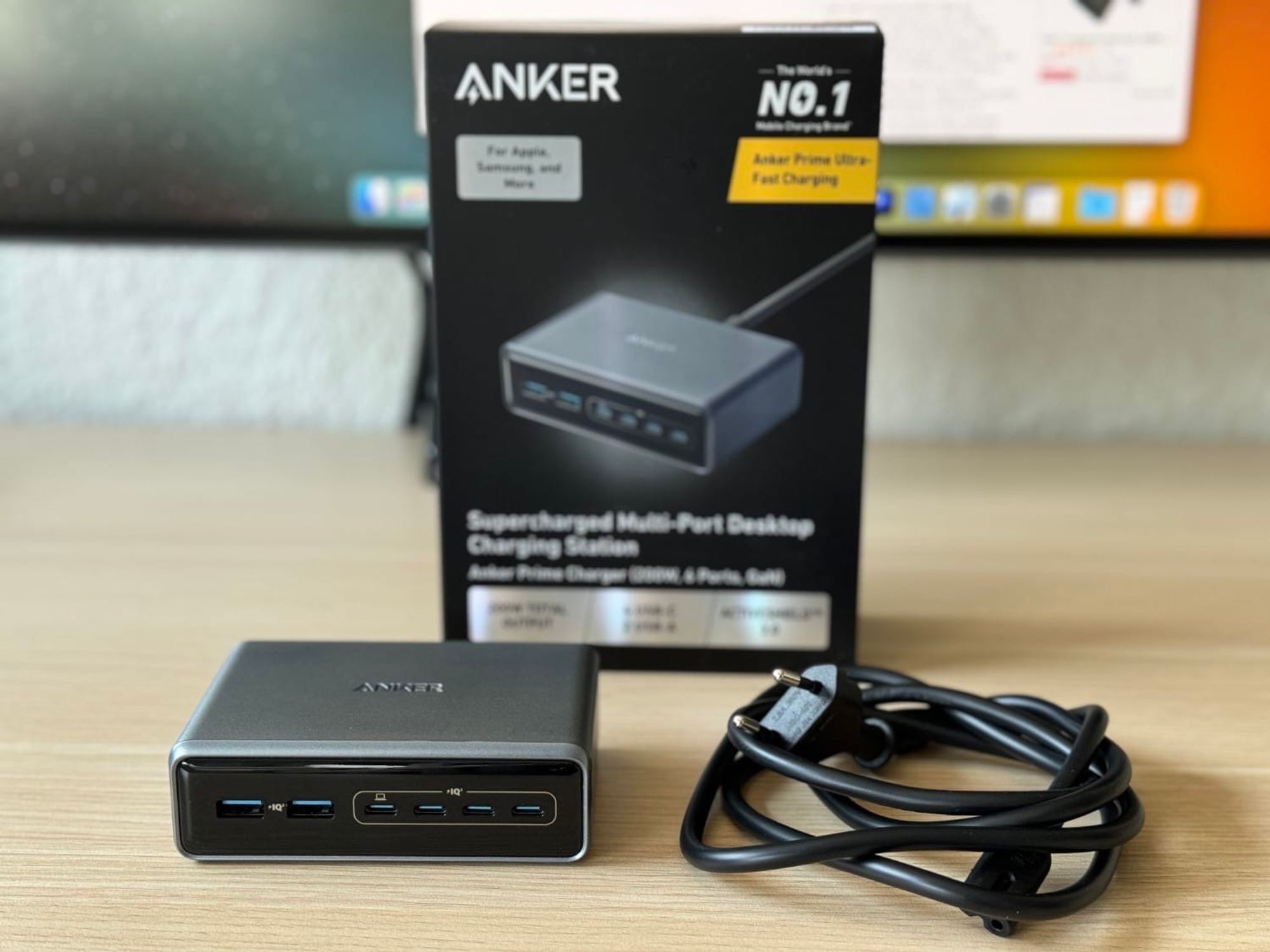 Das Anker Prime 200W Ladegerät liegt samt Stromkabel und Verpackung auf einem Schreibtisch, im Hintergrund unscharf ein Monitor mit macOS-Desktop. 