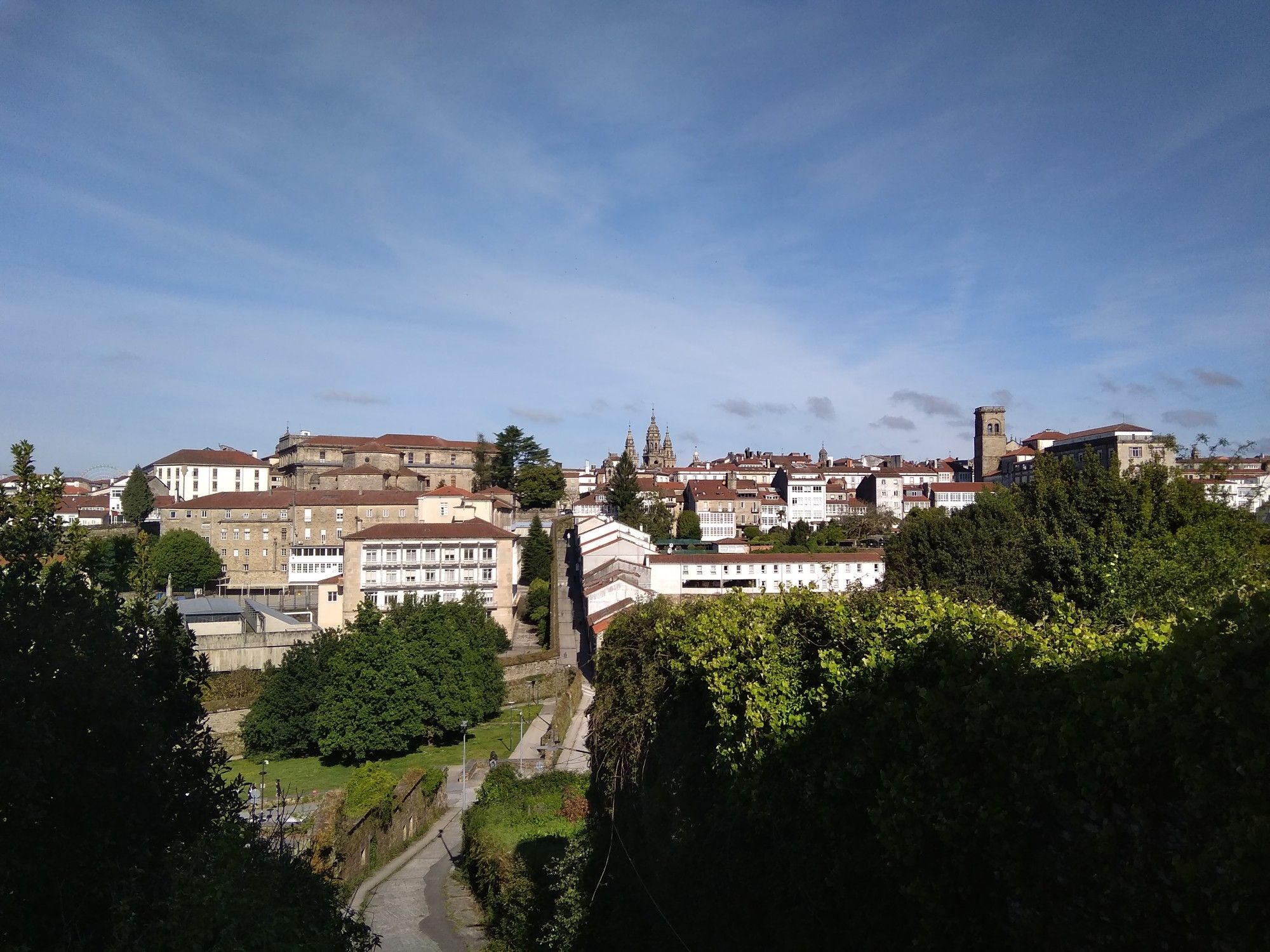 In the foreground are some shade-covered hedges looking out over a small dene. The dene is crossed by a paved road towards the other side, where houses of various time periods comprise the city centre of Santiago de Compostela. Various church towers can be seen against a blue sky partly veiled by skeins of white clouds.