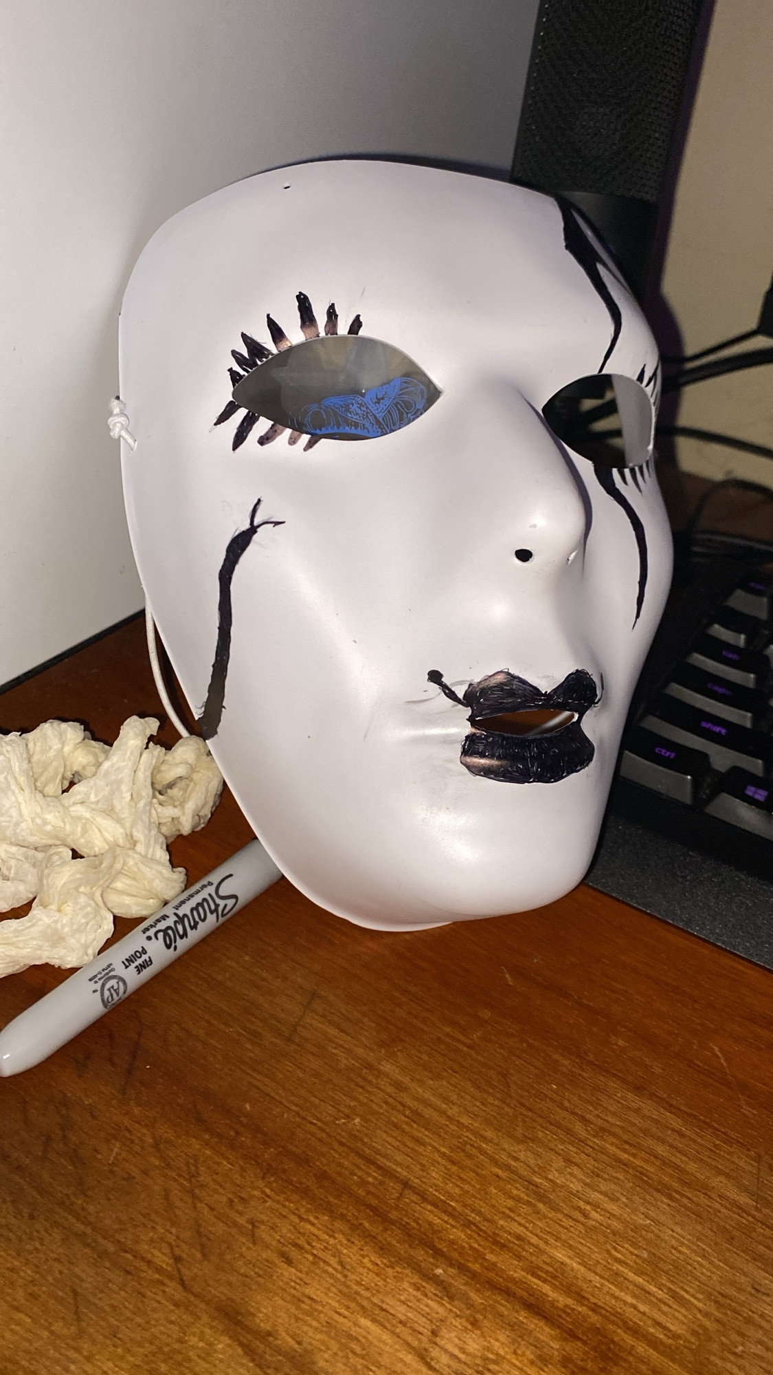 A white mask on a desk with black markings that look like cracks and eyelashes with lipstick, made to look like a porcelain doll mask