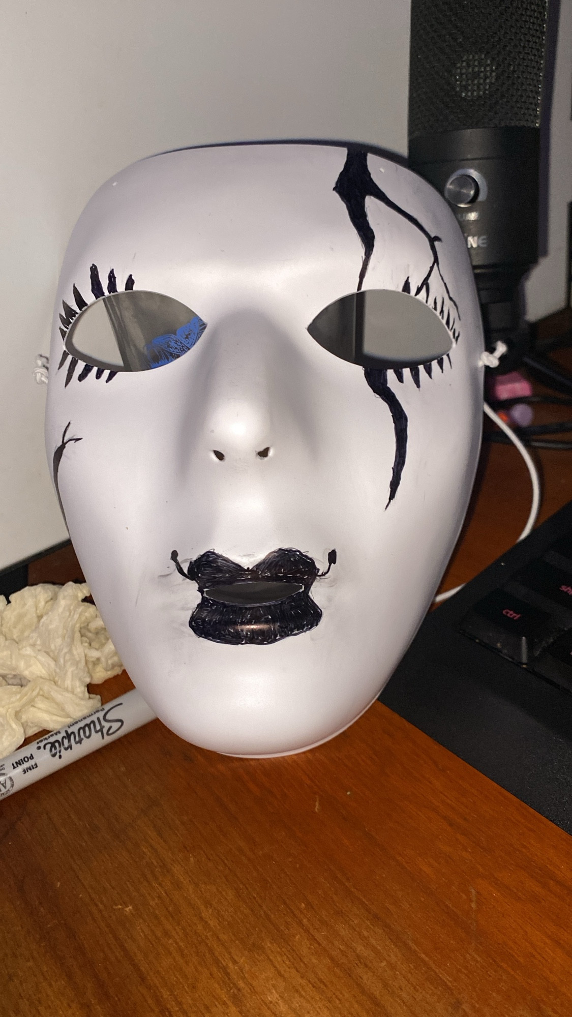 A white mask on a desk with black markings that look like cracks and eyelashes with lipstick, made to look like a porcelain doll mask