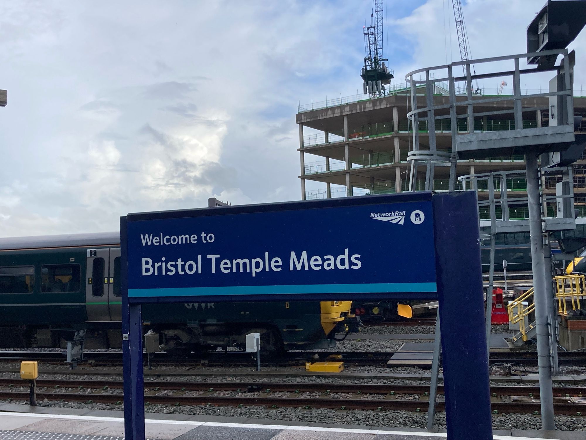 Ein blaues „welcome to Bristol Temple Meads“ Schild auf einem Bahnsteig. Dahinter Schienen, ein Waggon (links) und ein Gebäude im Bau (rechts).