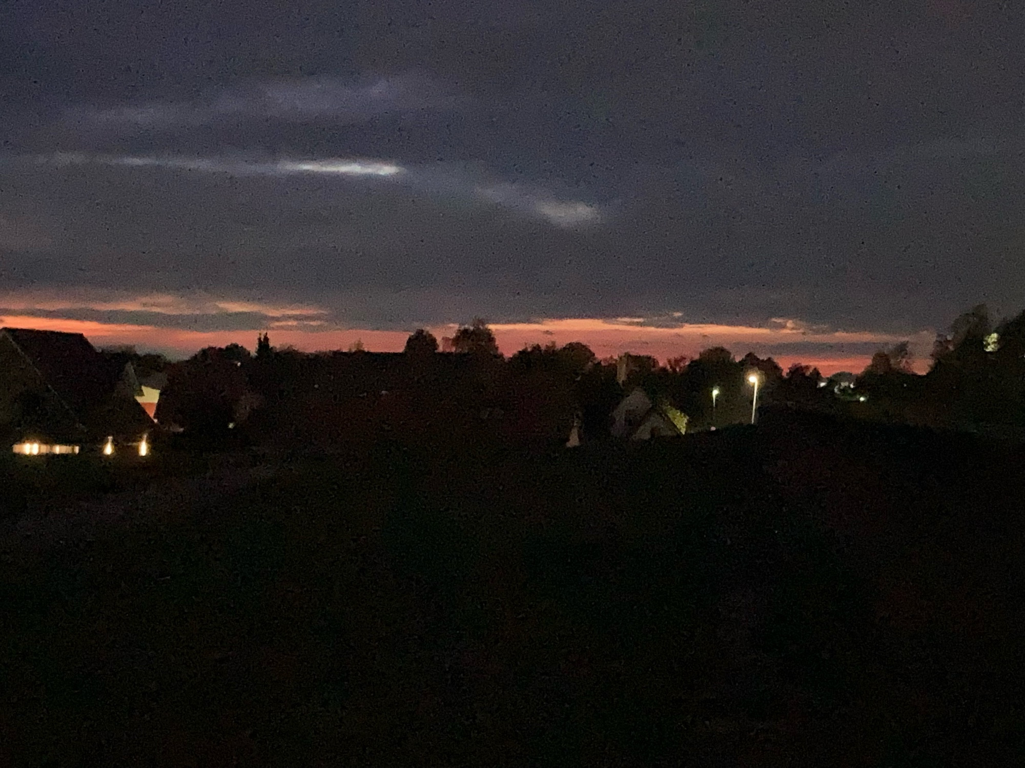 Sonnenuntergang. Die Landschaftssilhouette unten ist schwarz, Baumkrone und Hausdächer sind teilweise erkennbar, einige wenige Lichter links und rechts. Direkt über der Silhouette ein schmaler Streifen rotes Licht, darüber dunkles grau mit kleinen hellen Lichtstreifen links.