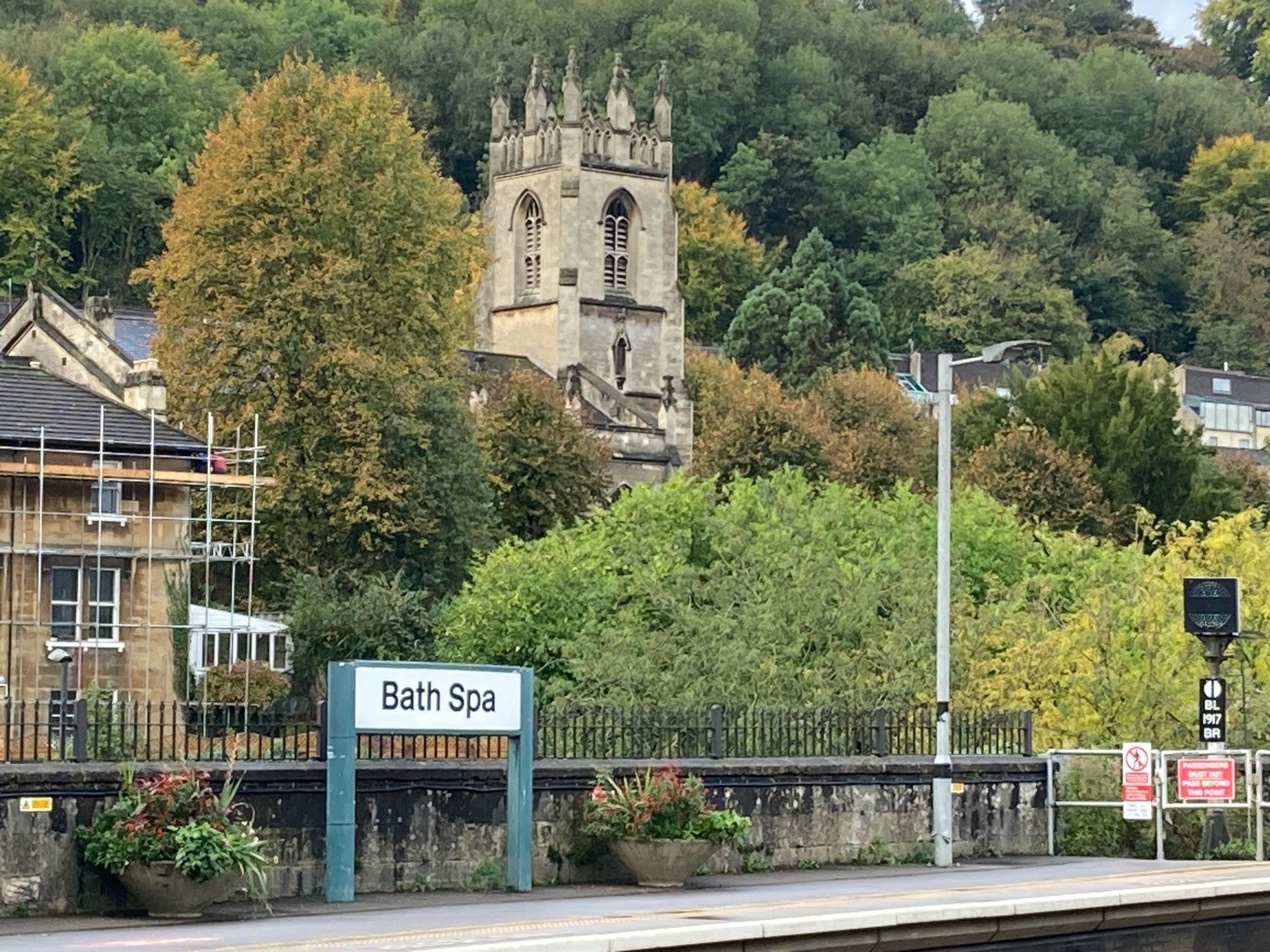 Wir schauen über Schienen auf den anderen Bahnsteig meist einem Schild „Bath Spa“ und dahinter Büsche, Bäume und ein Türmchen mit Zinnen. Das Bild ist nach oben mit Bäumen ausgefüllt.