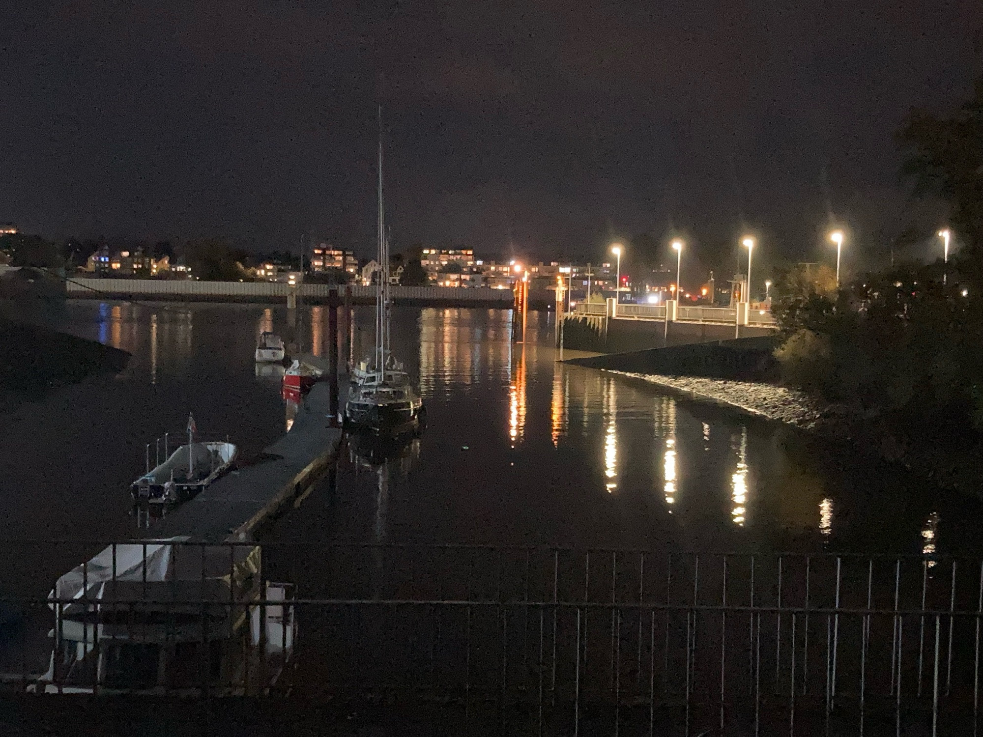 Blick über einen kleinen Hafen mit langem Steg und wenigen Booten in der  Mitte und einen quer dazu laufenden Fluss dahinter und das jenseitige Ufer. Am anderen Ufer sind viele bunte Lichter, die sich im Wasser spiegeln. Der Himmel ist dunkelgrau.