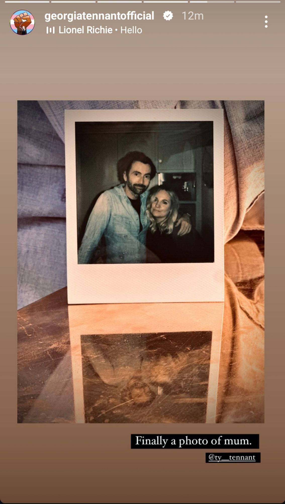 Screenshot of Georgia Tennant's Instagram story featuring a polaroid photo of David and Georgia Tennant. David has his arm around Georgia. Both are smiling and looking at the camera. The story is captioned, "Finally a photo of mum." Photo credit to Ty Tennant, with "Hello" by Lionel Richie as the background music.