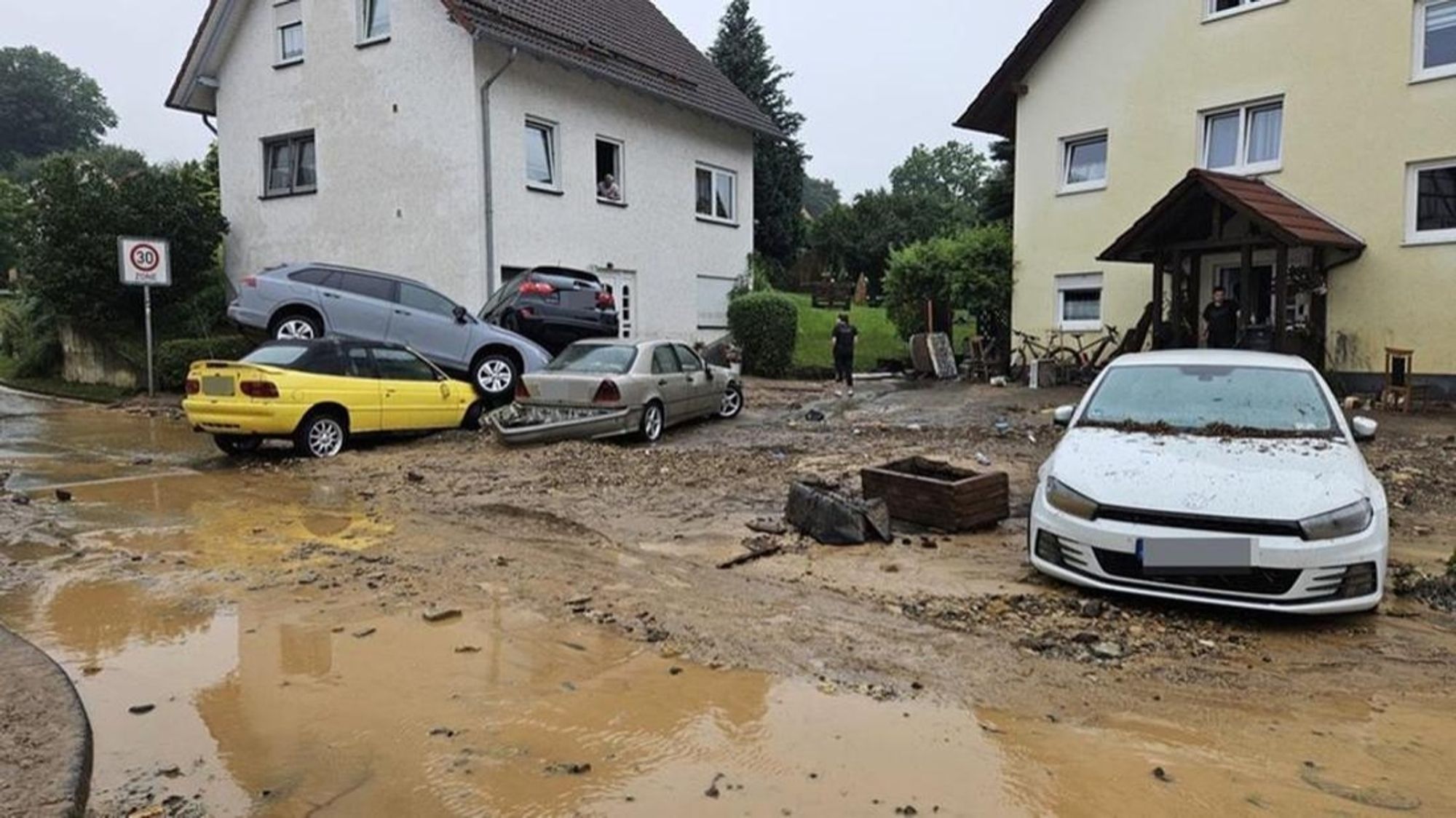 Im Hintergrund zwei Häuser im Vordergrund Matsch und Schlamm auf einer Straße und übereinander gestapelte Autos