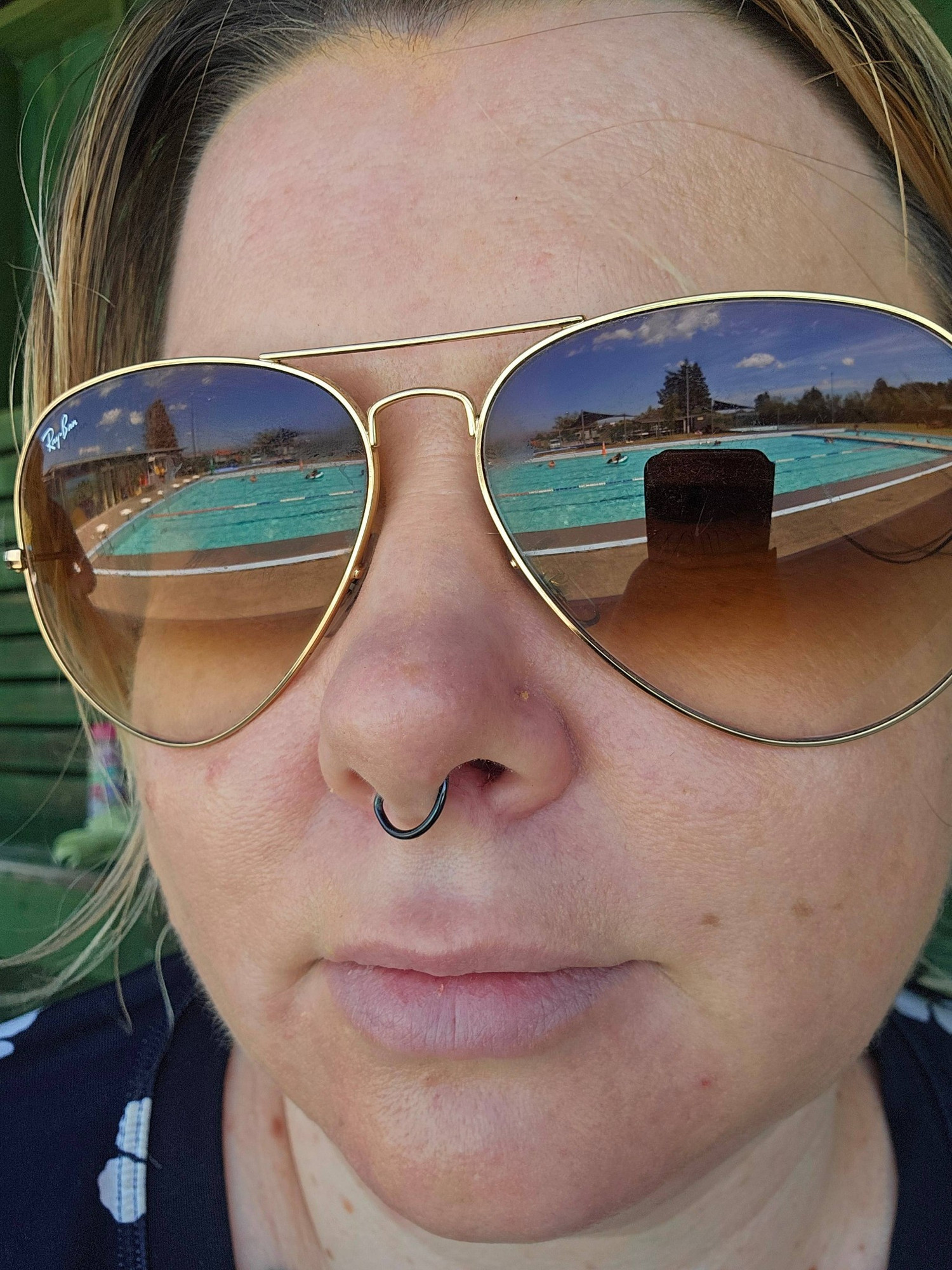 Woman wearing aviator sunglasses. The sunglasses show a reflection of a large outdoor pool