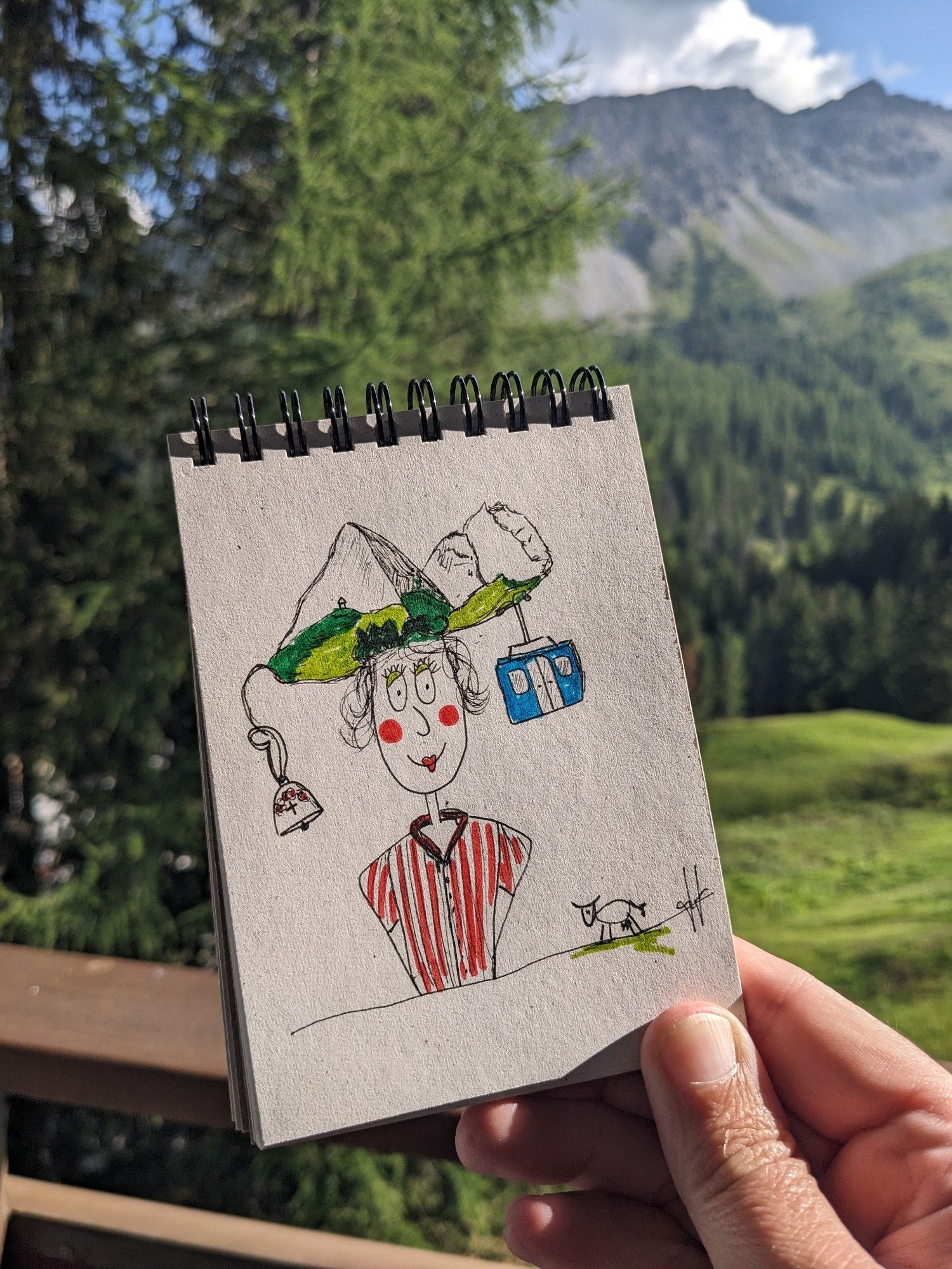 Foto vom Skizzenblock. Im Hintergrund Wiese und Berg. Auf dem Papier Portrait einer Fantasyperson mit Bergen in den Haaren und Seilbahngondel am Berg.