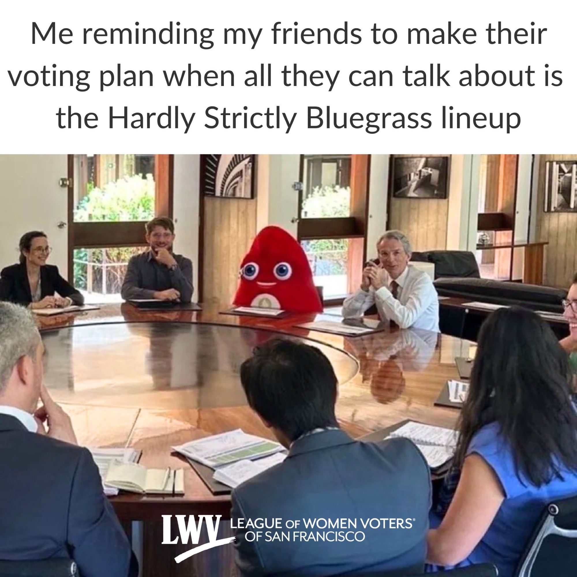 A group of people sitting around a large round meeting table talking to each other. Sitting in one of the chairs is the cute mascot for the Olympics called Phryge. It is a human-sized red stuffed plush figure with vacant wide eyes and smile. None of the people sitting at the meeting table is acting like it's weird that the cute mascot is at the meeting. Even though it's smiling, in this scene the mascot looks hilariously like it doesn't belong. Above the image, the text says "Me reminding my friends to make their voting plan when all they can talk about is the Hardly Strictly Bluegrass lineup"