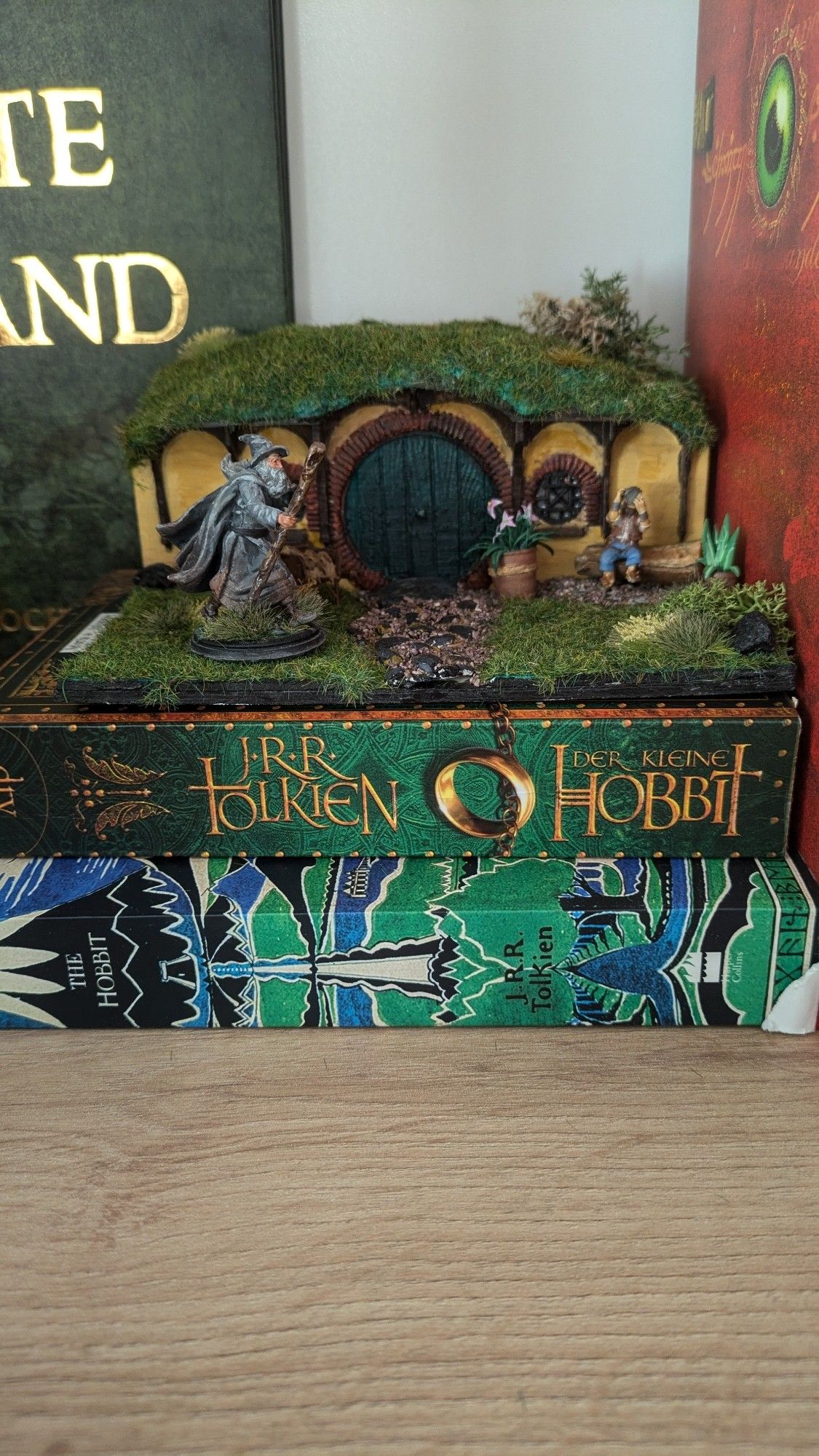 Auf dem Buch "Der Hobbit" von Tolkien (in deutscher und englischer Ausgabe) ist ein Miniaturmodell einer Hobbithöhle vor der ein Hobbit auf einer Bank sitzt. Gandalf geht auf dem Rasen spazieren. Das Modell ist nicht breiter als das Buch (also rund 20cm) und etwa 8cm hoch.