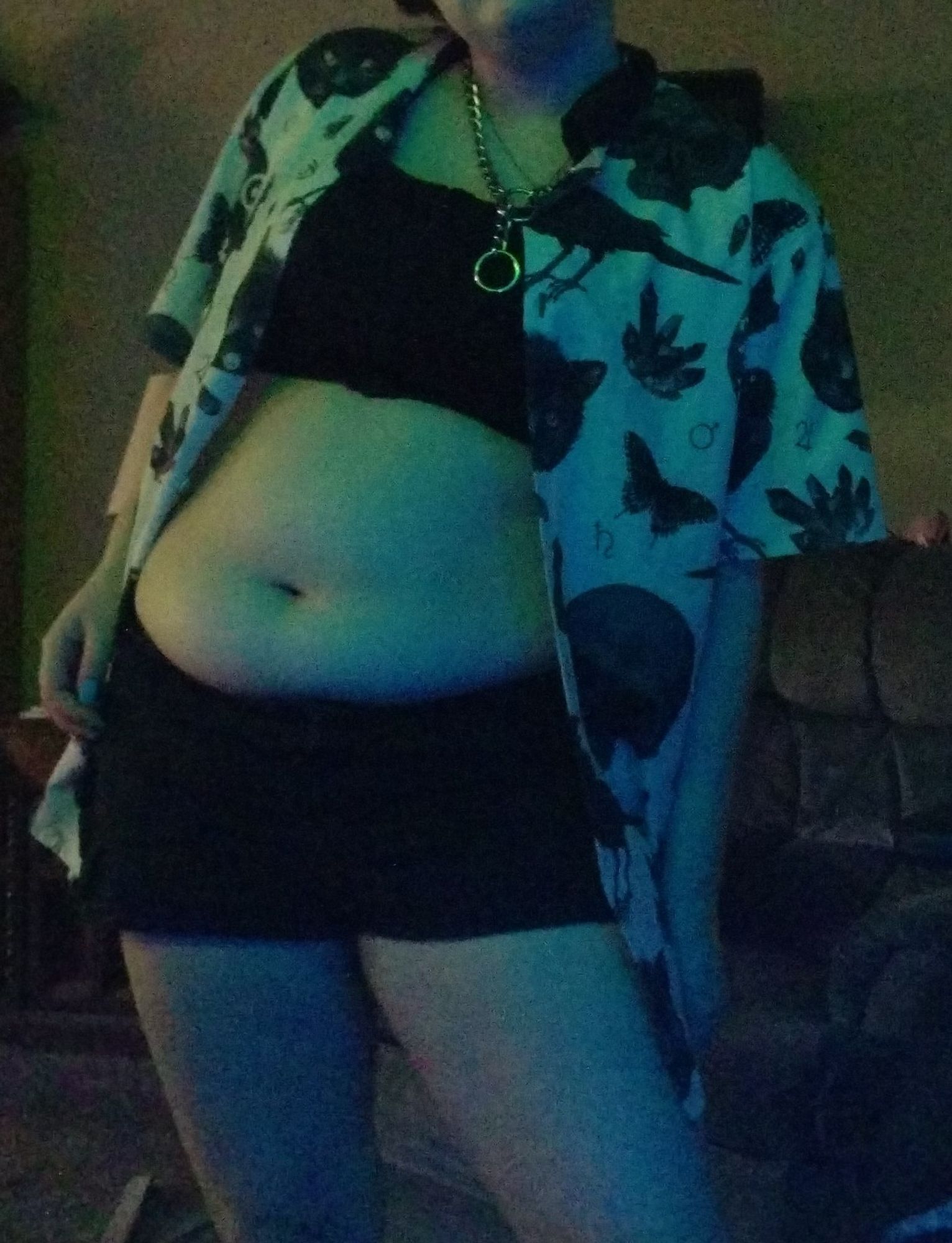 A photo of me, a slightly chubby Demifluid 19 yo, afab, I'm wearing black shorts, and a black sports bra, with a various witchy patterned button up T-shirt which I have undone. I'm also wearing a choke chain loosely around my neck. The shading is blue, while the lighting is green. I'm standing at about 5'4.