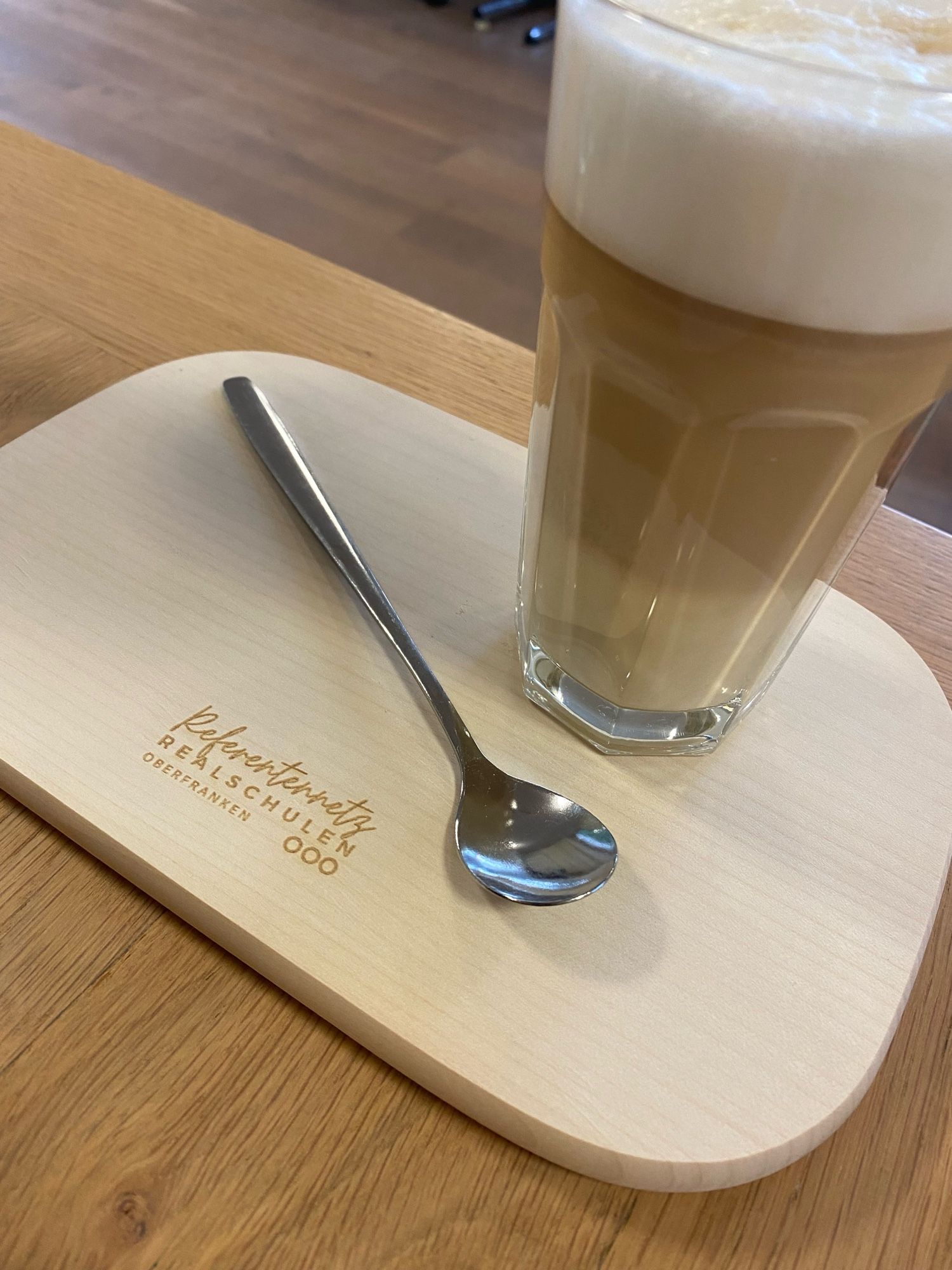 Picture of a glass of coffee, a spoon and a wooden plate with an engraving that says „Referentennetz Realschule Oberfranken“