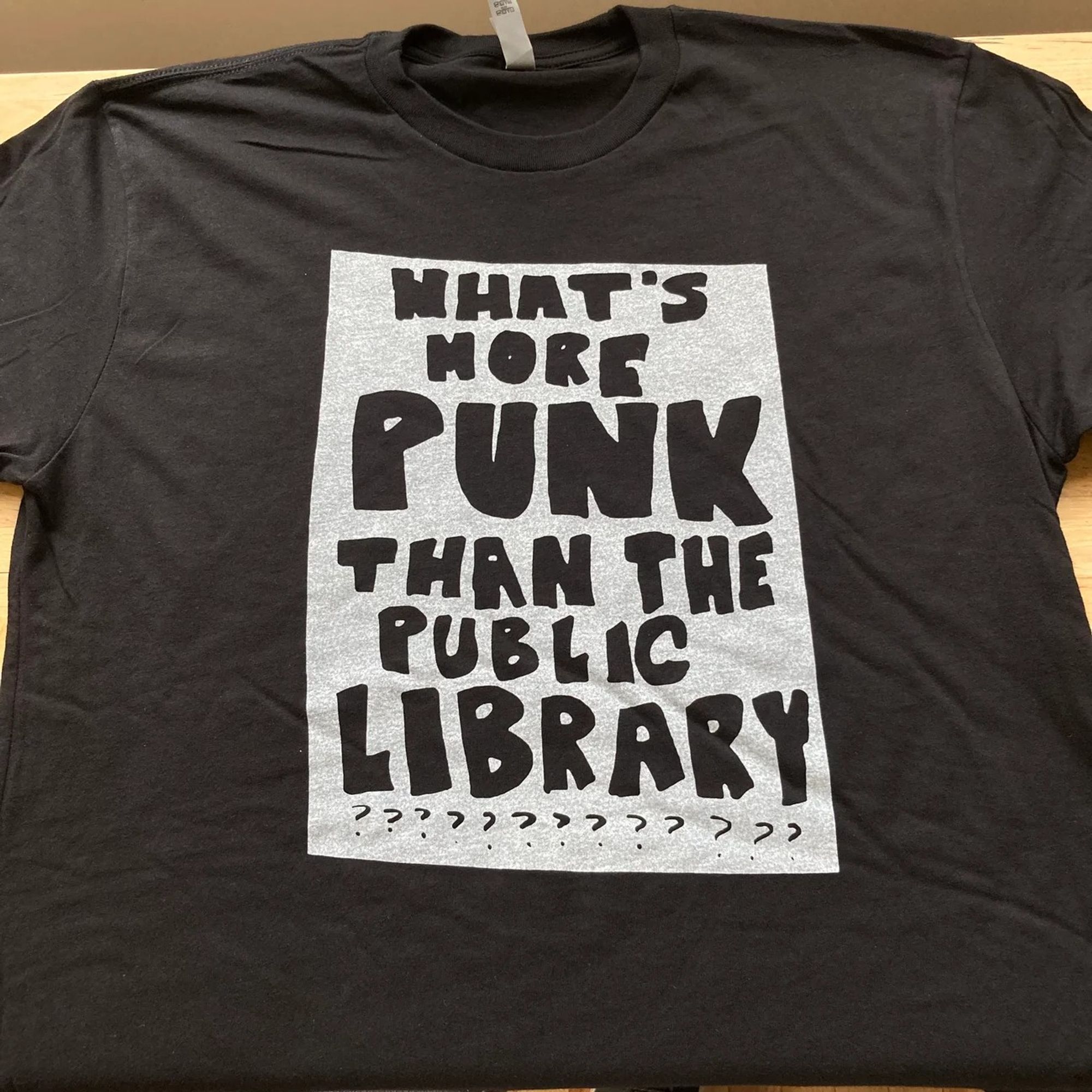 Black t-shirt with a white silk screen block. In "punk" style letters in black (not silk screened, they're parts of the t-shirt that were left without screening) the words What's more punk than the public library??????