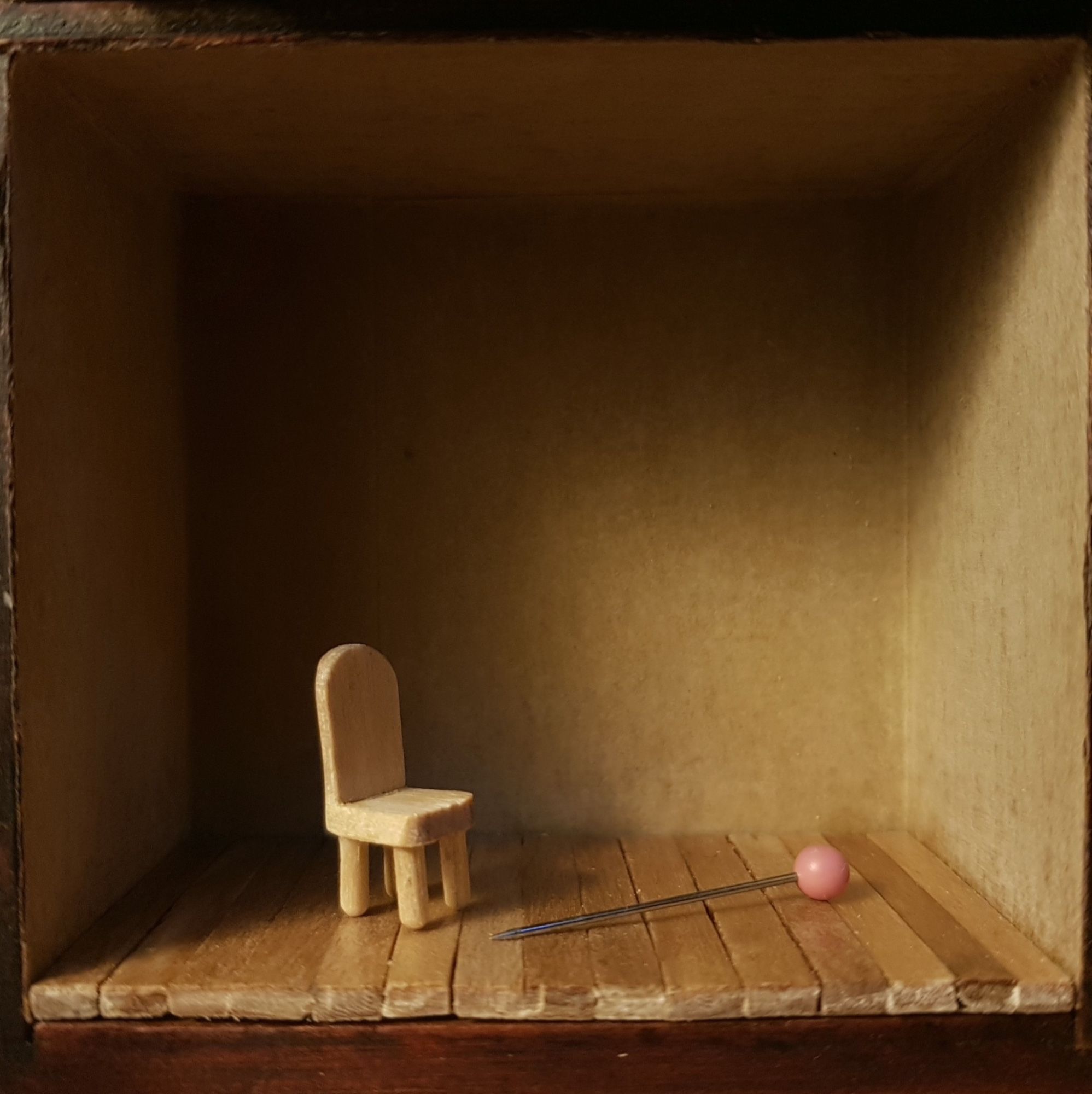 Tiny wood chair on tiny wood floor next to a pink headpin in a box.