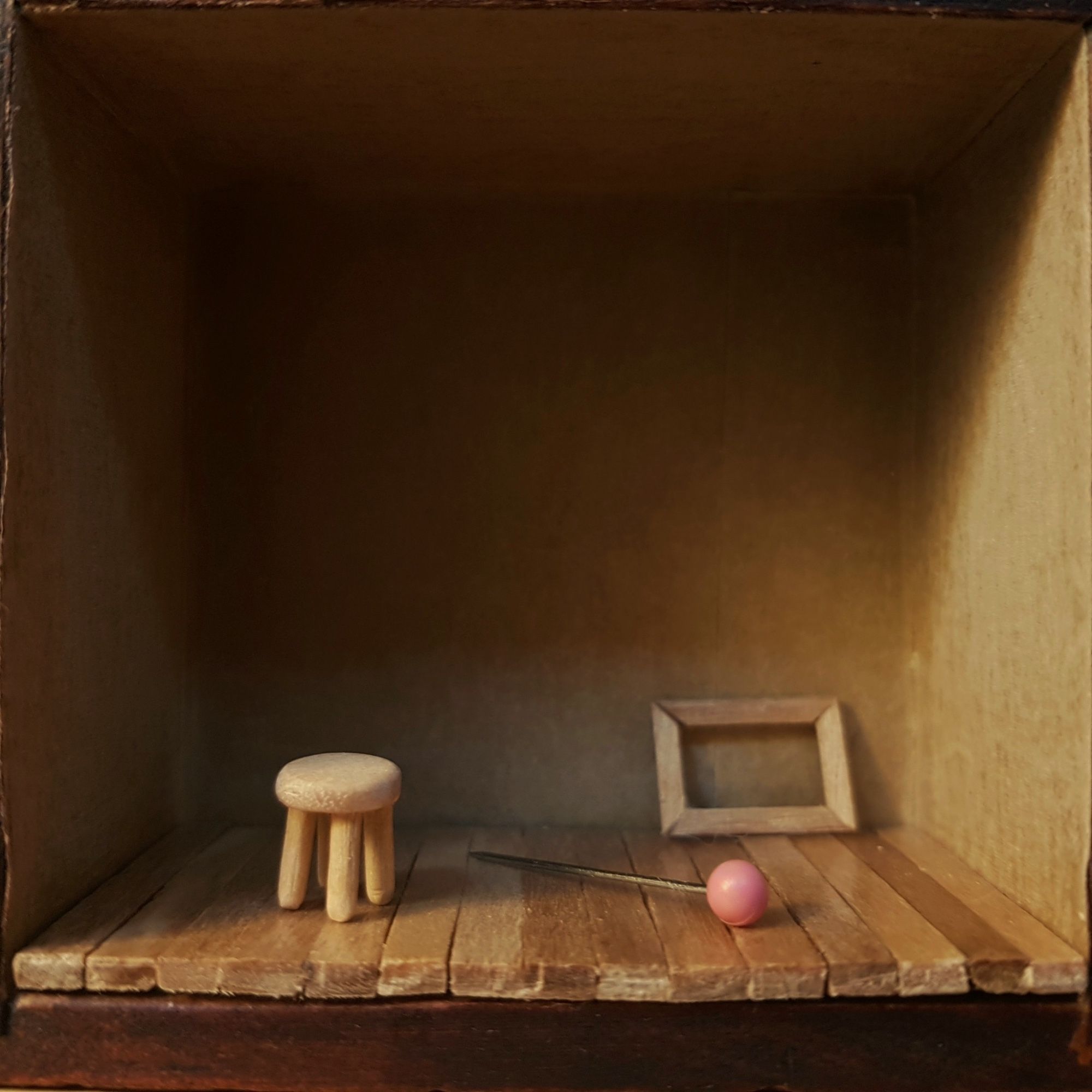 A miniature room with a tiny wood stool next to a pink headed pin and a tiny empty frame leaning against the wall on wood floor