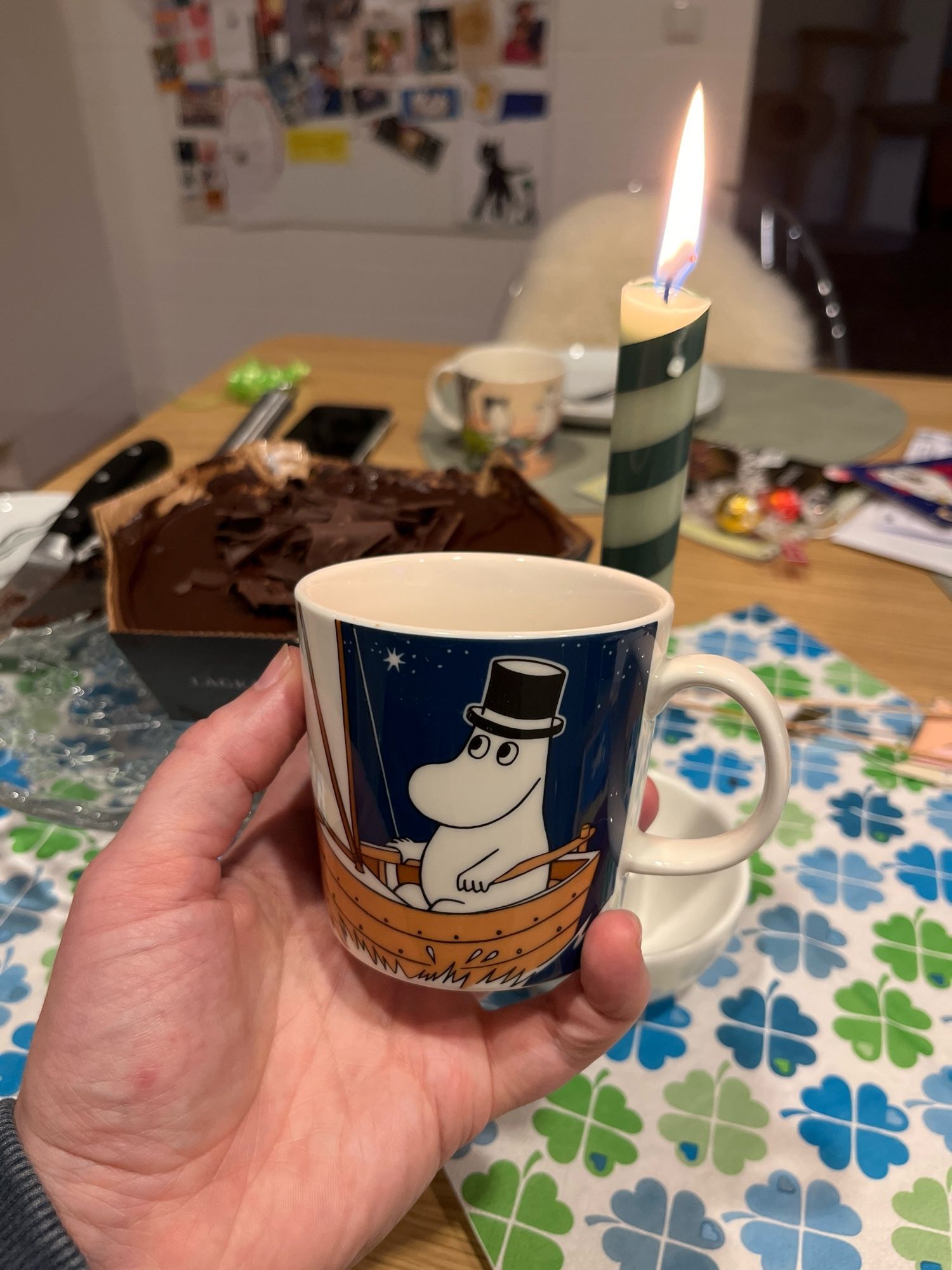 Foto einer Moomin-Kaffetasse, einer Kerze und im Hintergrund Kuchen und etwas Deko