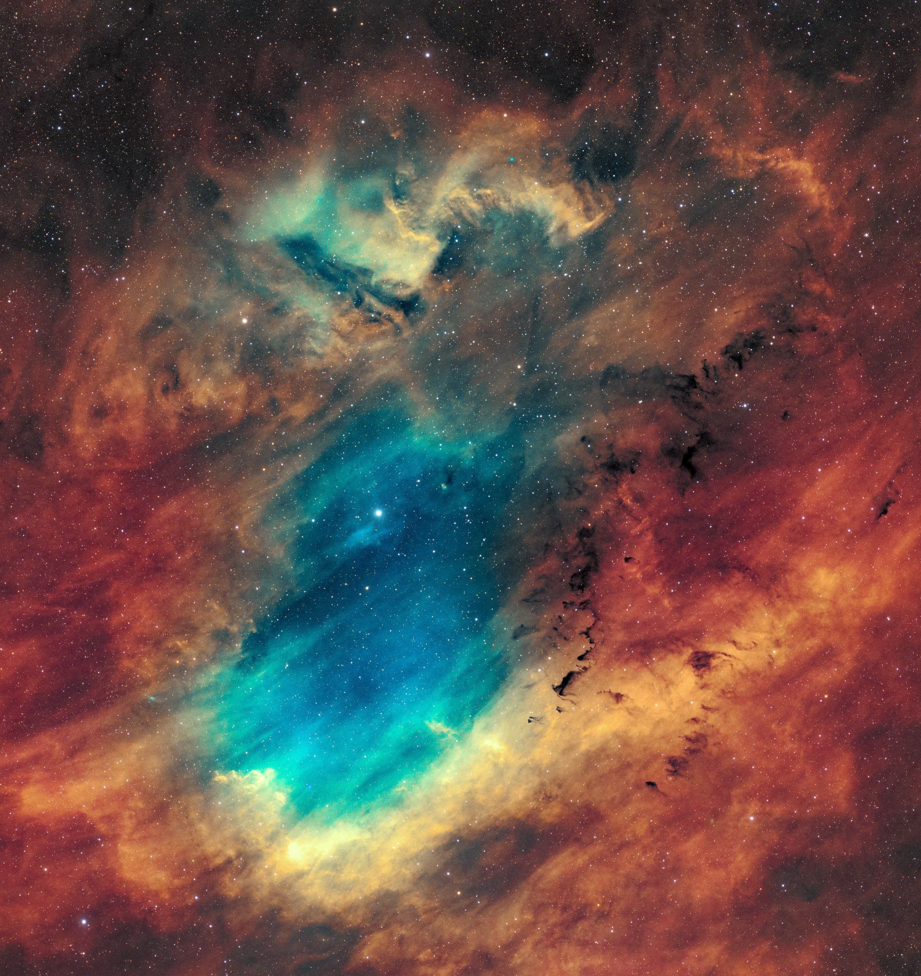 A sort of bean shaped colorful and bright space nebula, a blue and teal center surrounded by reds and oranges showing the various gasses the nebula is composed of. The gasses have a very wavy structure and almost look like a beach front or atoll. There seems to be diagonal movement as well. Some smaller wispy dark cloud structures are visible on the right side of the nebula. There's a myriad of stars.