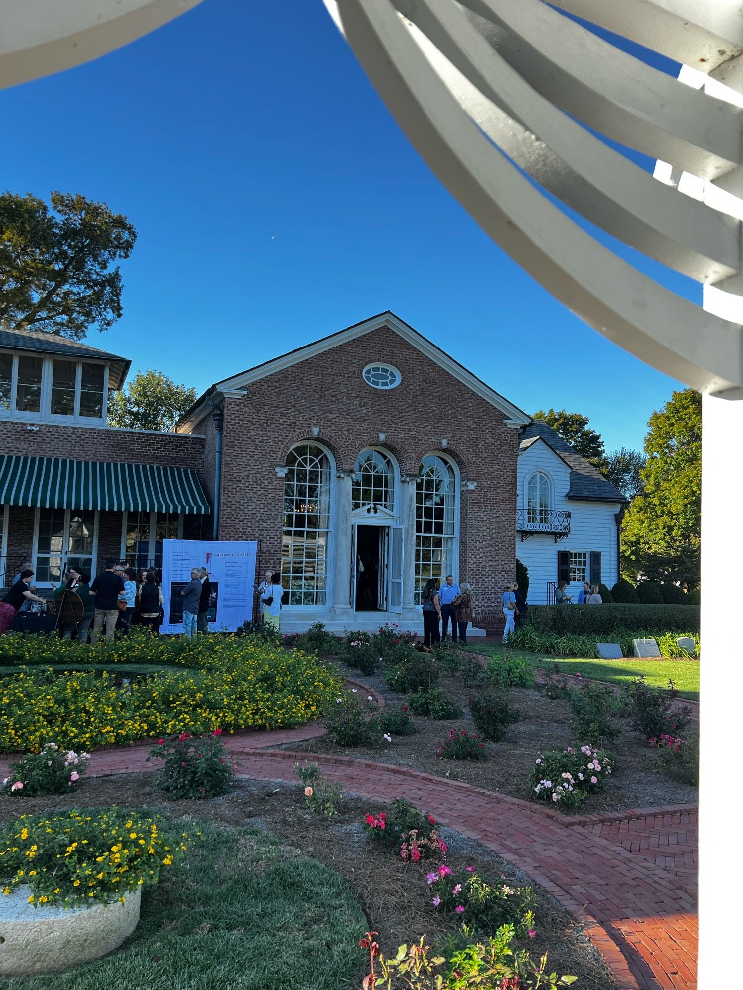 Photo taken from the gazebo with the gazebo doorway framing the large building with a lot of people and a lovely garden and pond.