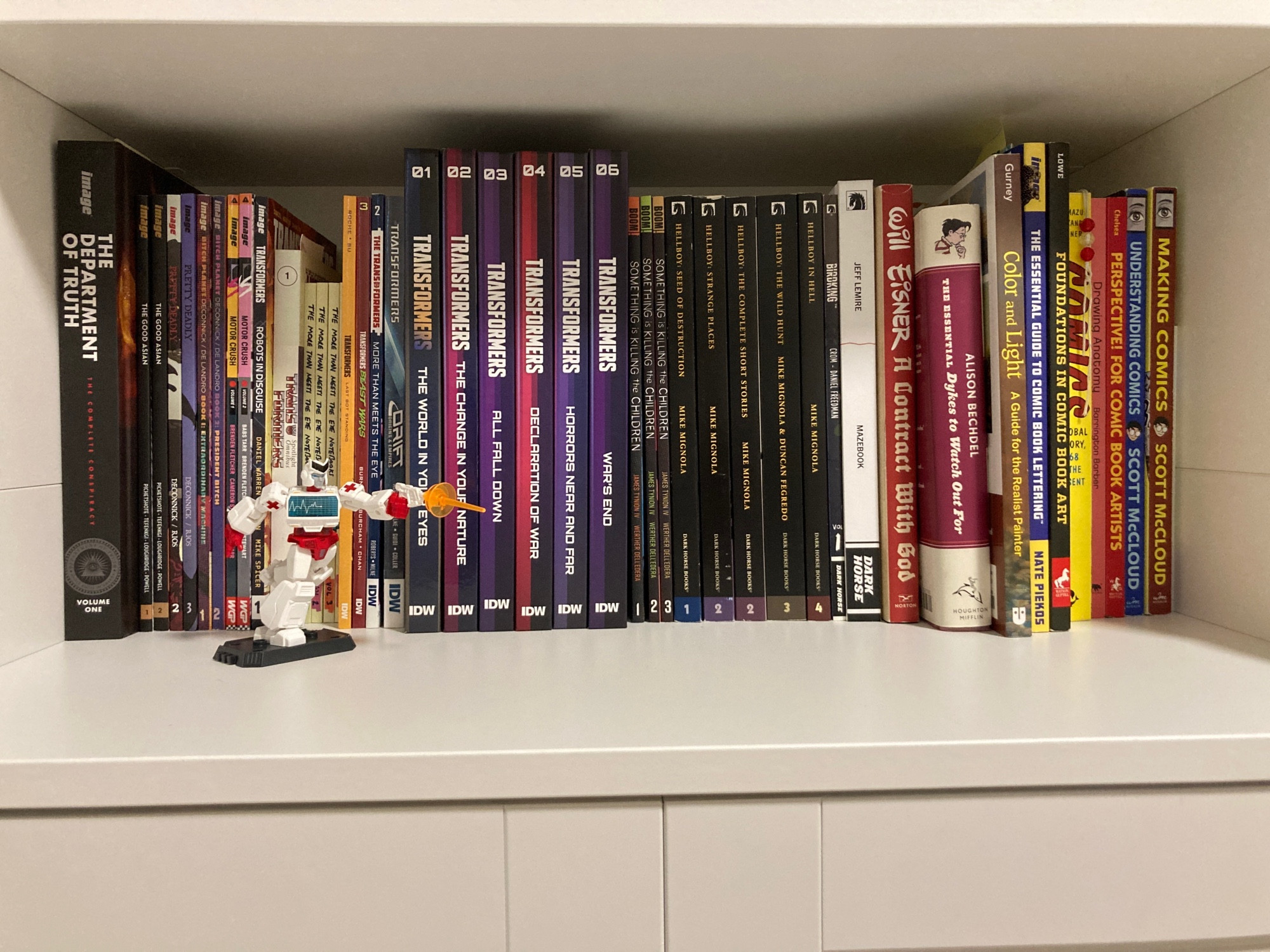 A shelf of comic books including the Department of Truth, The Good Asian, Pretty Deadly, Bitch Planet, Motor Crush, a variety of Transformers series, Hellboy, Something is Killing the Children, Birdking, Mazebook, A Contract with God, Dykes to Watch Out For, a variety of comic and art theory textbooks, and a little figurine of the transformer Ratchet.