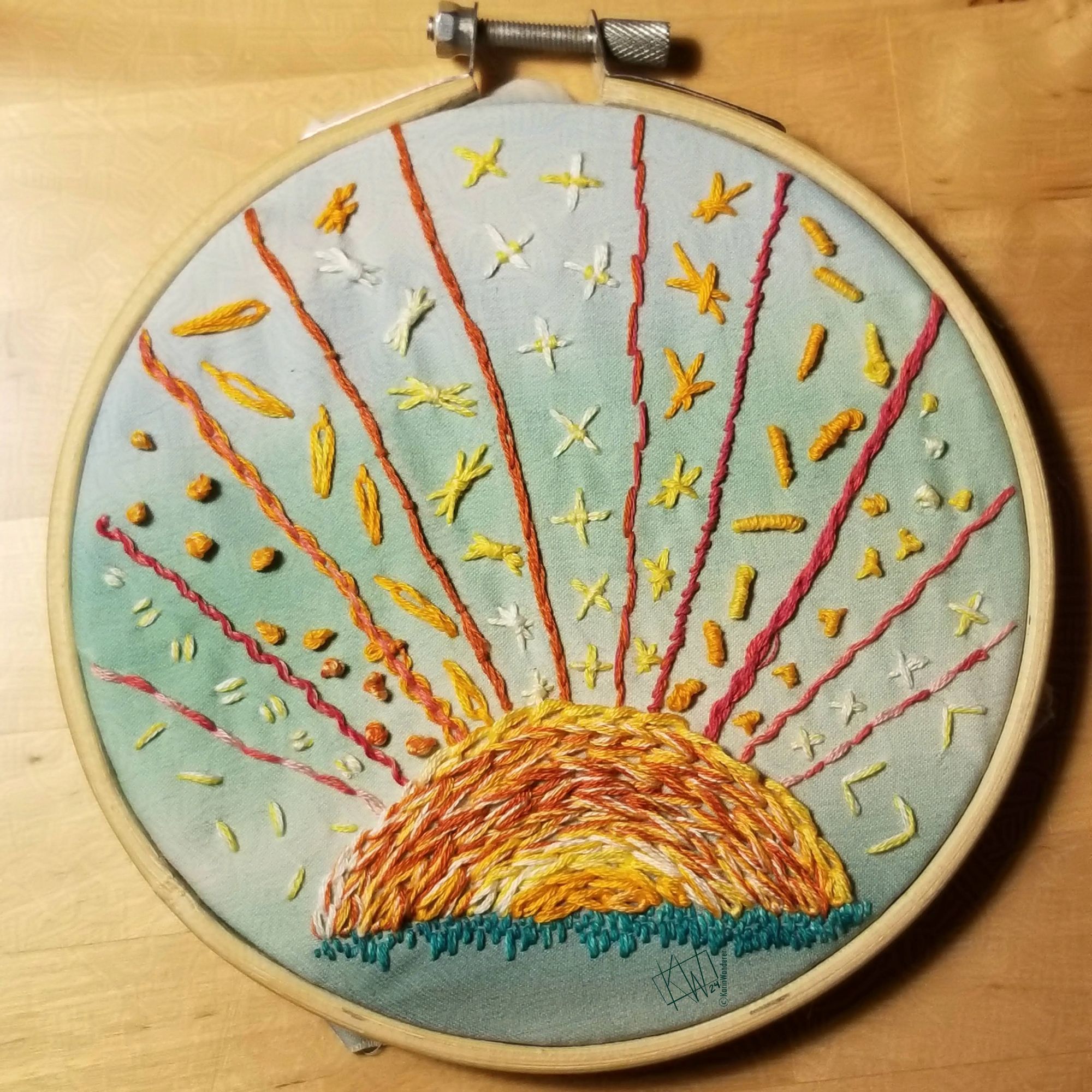 Embroidery of a setting sun with rays in different embroidery stitches in yellow, orange, & pink.