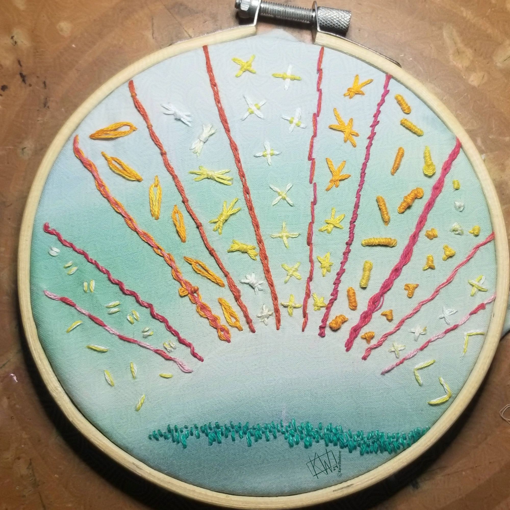 A mostly-finished embroidery hoop of a setting sun with rays in different embroidery stitches in yellow, orange, & pink. The sun has not been stitched yet.