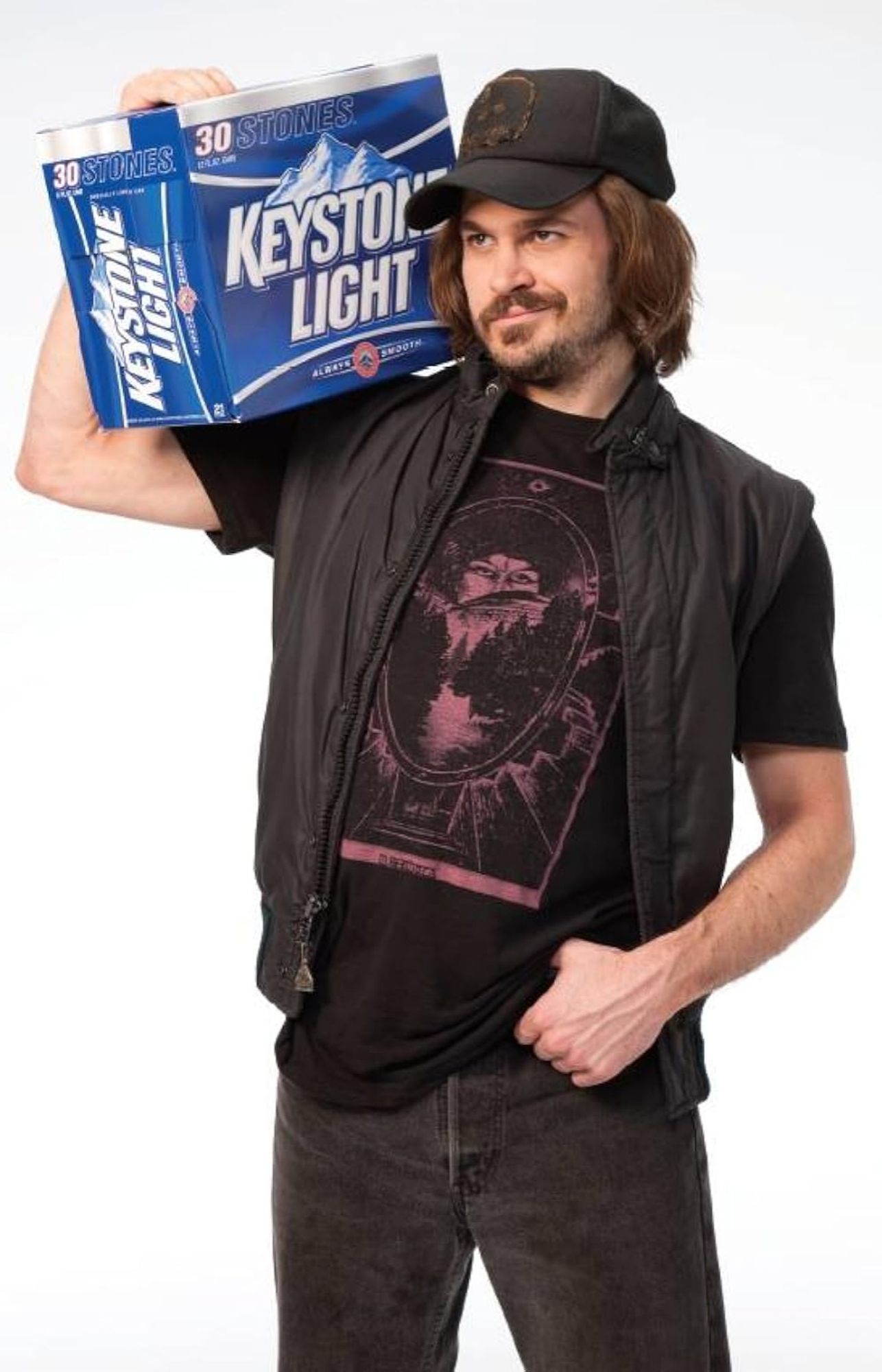 A picture of a white man wearing black pants, a black T-shirt and a black vest holding a 30 pack of Keystone Light on his shoulder like a boom box.