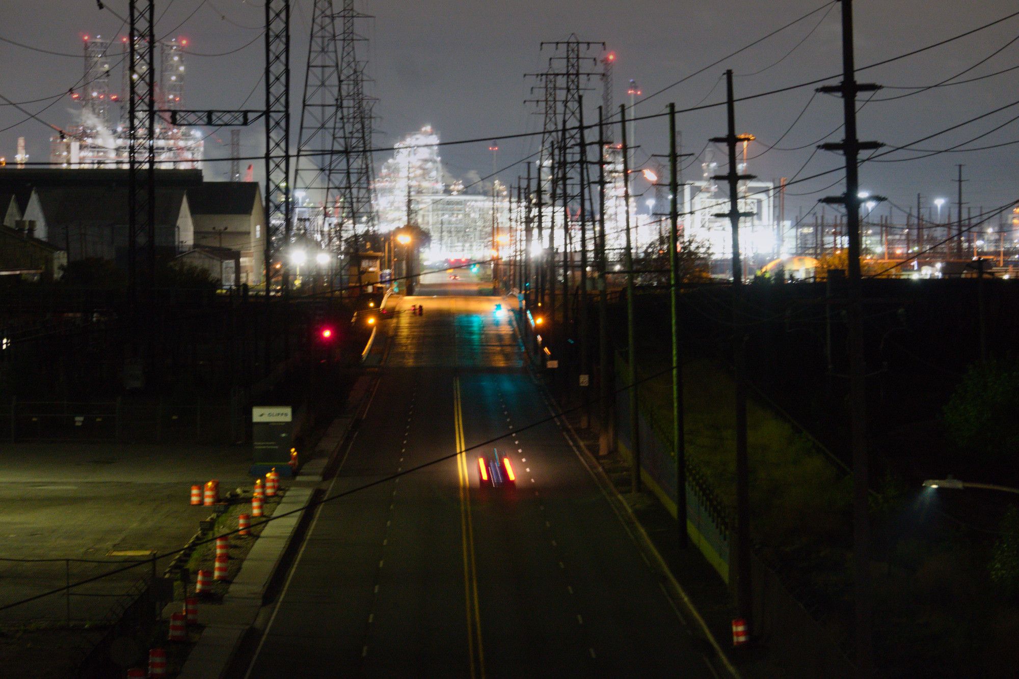 We're on an elevated toll road, standing on the shoulder, looking northeast up Dickey Rd, which has a little hump of a bridge that crosses the Indiana Harbor and Ship Canal, then approaches BP's whiting refinery, which is bright white contrasting against a nearly dark night scene in the rest of the image. A lone car races away from us on the road, leaving a blur of brake lights. Along the left side of the road, we see some leading lines of orange cones pointing up toward the horizon. Between the cones and the refinery is Marktown. On both sides of Dickey Rd, tall pylons carry industrial-grade electricity to the refinery, and to the sprawling industrial hellscape surrounding it on all sides.