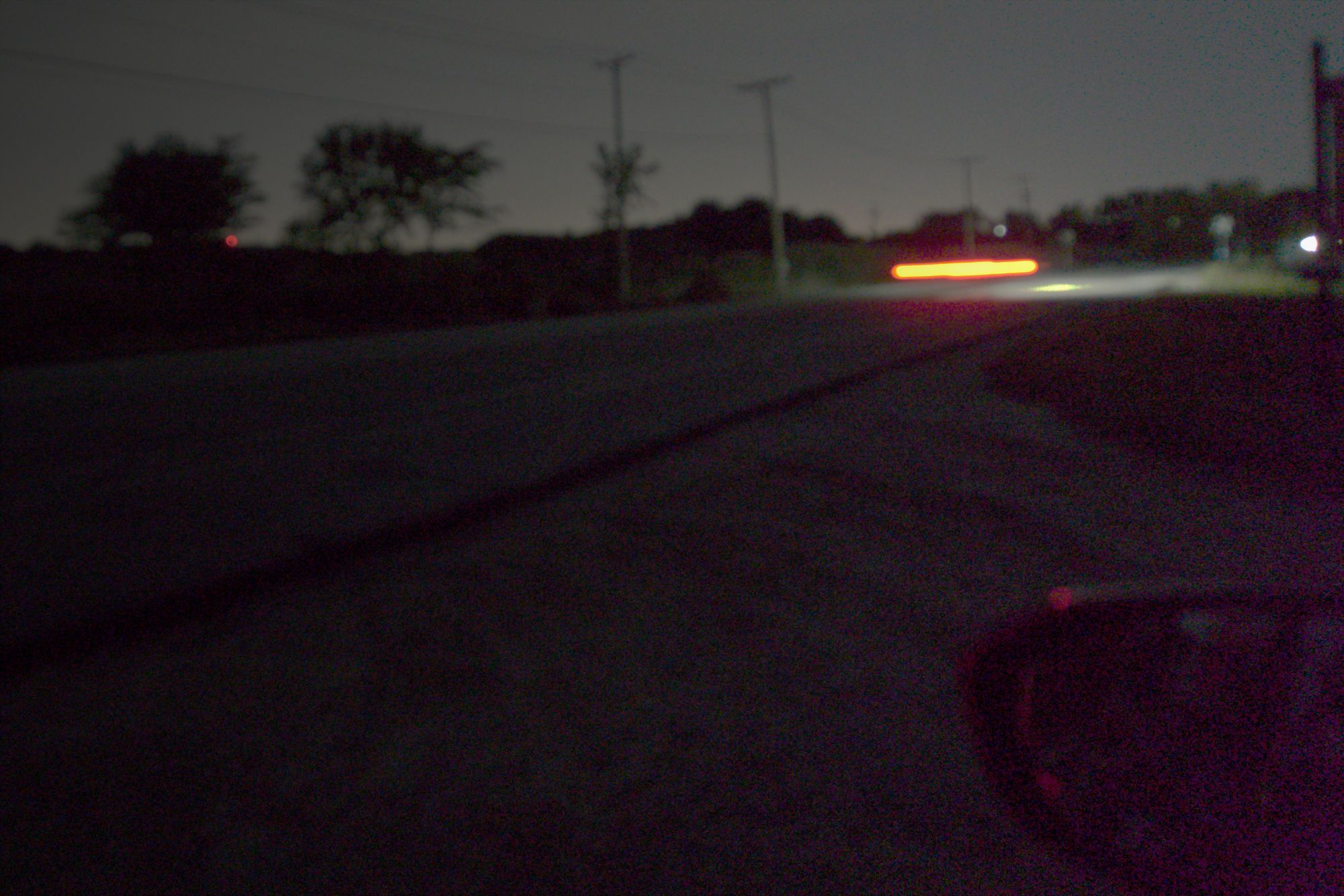 We're in a black car, looking out the window, in a fairly desolate location. It's night and there is hardly a streetlight in sight. Perfect location for mischief. The road extends out away from us to the right upper corner of the frame. On teh far side we see a few small trees, some power or telephone lines, and a mysterious red flash.