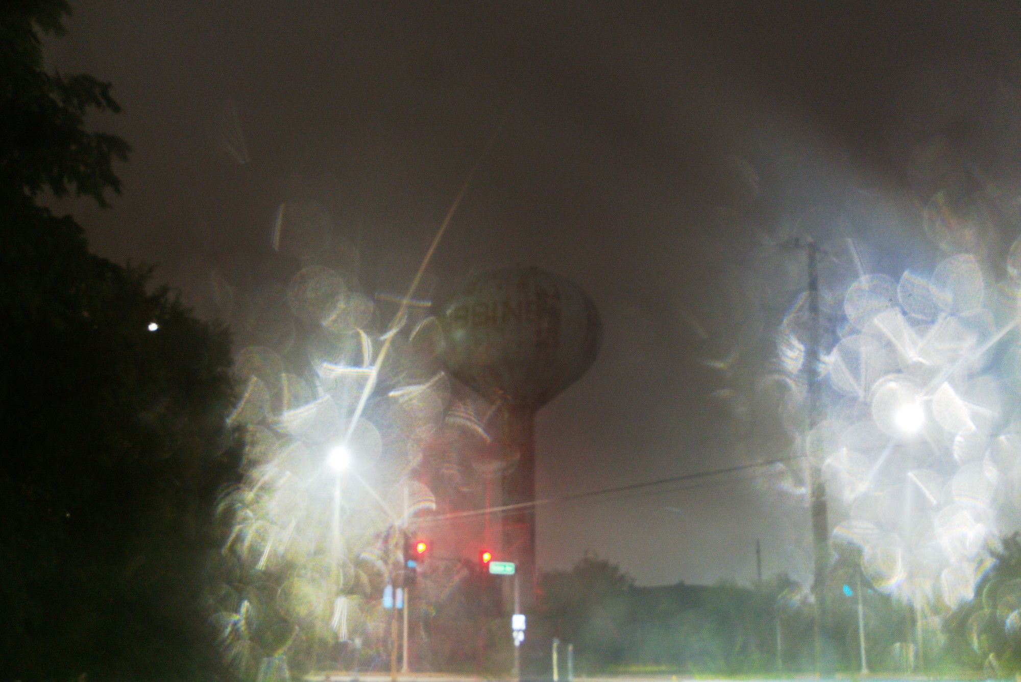 This is a very dark and murky photo. The main element is a small town's water tower, which is barely visble in the dark. In the foreground, two streetlights dominate the photo because water on the lens is scattering the white lights in dramatic and chaotic ways.