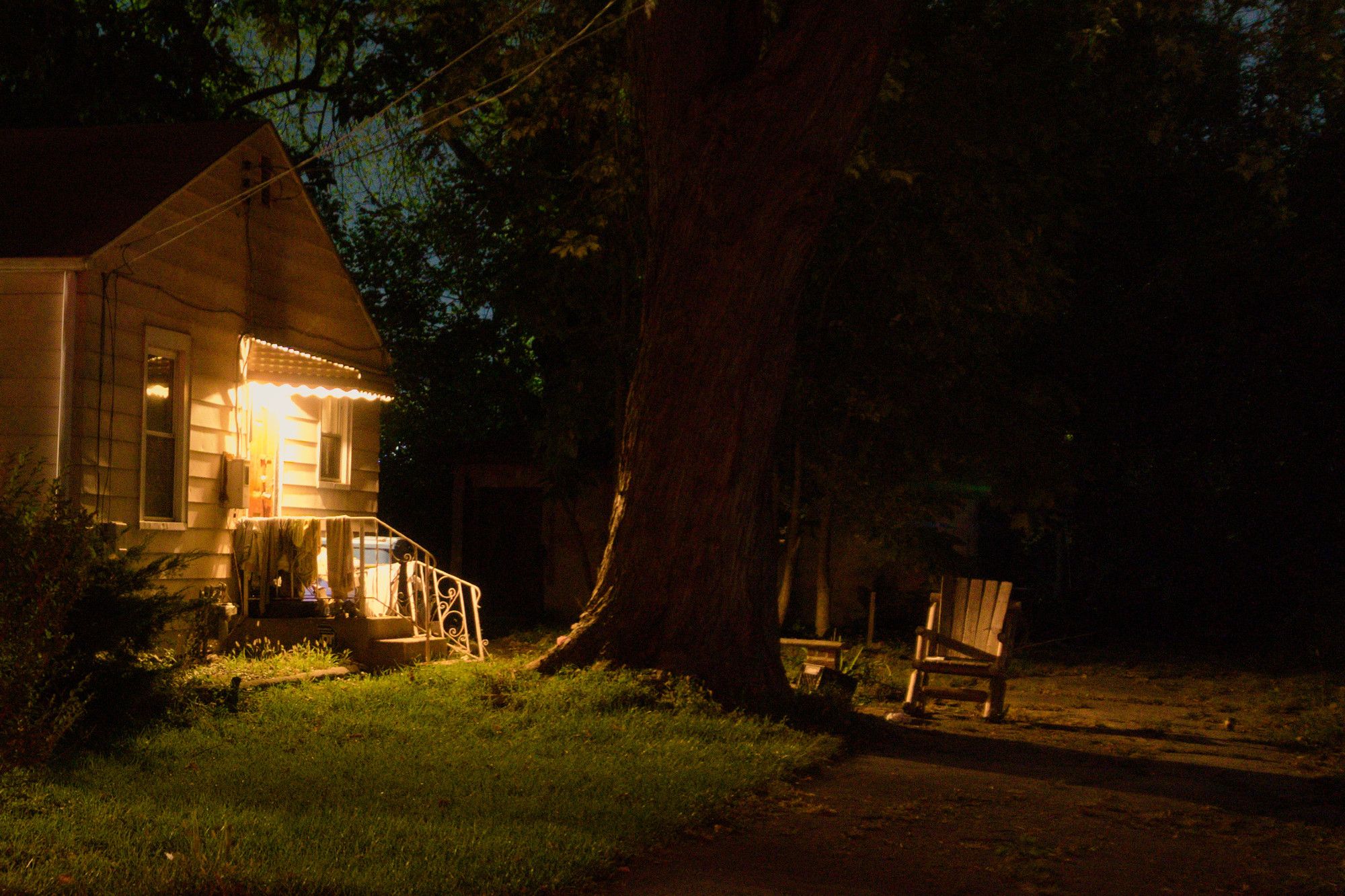 We're in the street, looking at the side lot next to a one story home. It's night and the scene is mostly shadows. On the left, the white/beige home has a yellowish warm lone light illuminating the scene from the side entrance to the home. That home is up two to three concrete steps. The right railing of that stairway is broken and leaned up against the steps. There's a really large tree in the center of the image, it's trunk perhaps big enough that two adults would be needed to wrap their arms all the way around it. On the far side of the tree, an Adirondack chair sits, lit in high-contrast, warm light. It's a little bit eerie, a little bit forlorn, a little bit inviting, a place you might want to escape and sit for a bit to think, but while also wondering who's watching, from the shadows, or -- from the car where I sit, taking photos.