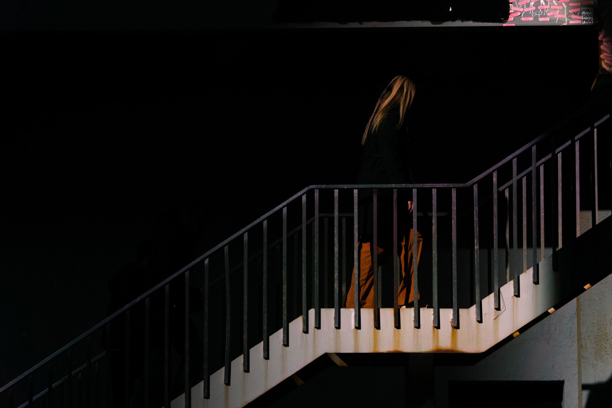 A staircase enters the frame at the bottom-left corner. It ascends for a bit, then levels out, then ascends up and out the right side of the frame. A woman is on the flat part walking to the right. The scene is very dark, and so most of the space is black. The woman's burgandy pants and reddish hair are visible, but not much else. Similar tones are in rust on the stairway below her, and in red graffiti above her. The whole shot is almost solely black, white, and splashes of red.