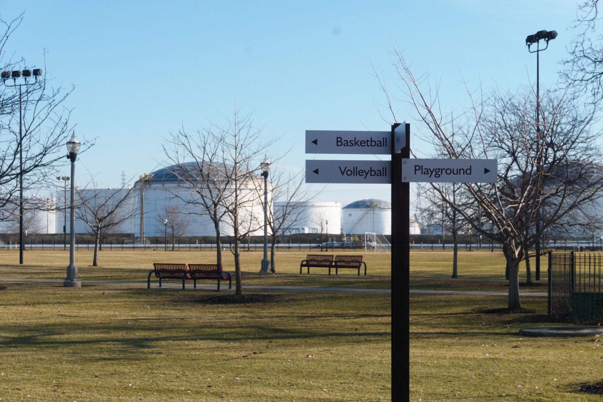 Here we're standing in a field. In the foreground is a pole with signs pointing left and right reading "basketball", "volleyball", "playground". But given the composition of the image, the signs are pointed at these same white petrochemical containers. But they're not even the SAME ones. There are several dozen, and despite looking identical, these are different ones from the last frame.