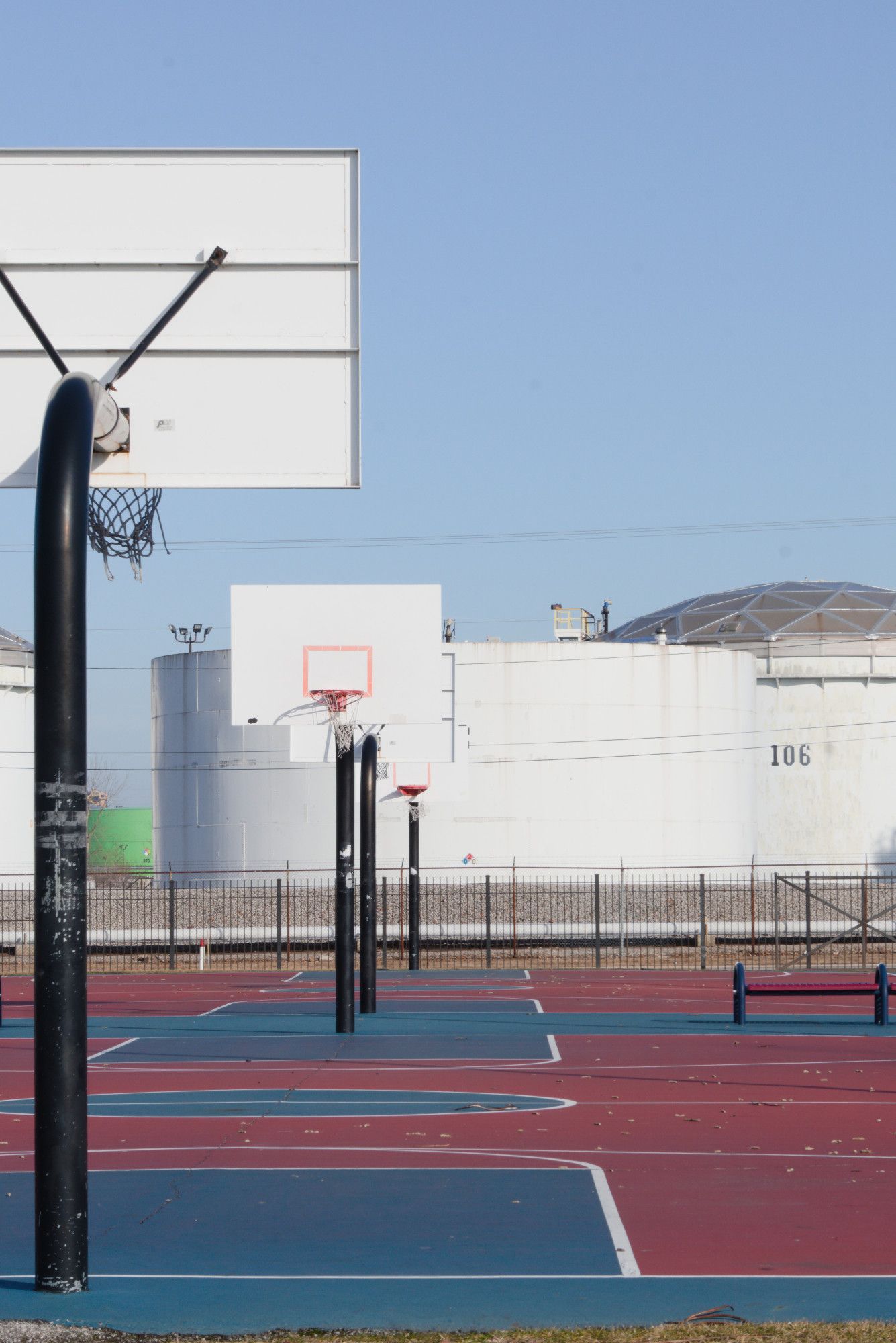 We're standing on a series of basketball court. We're looking from behind the first backboard, down across two courts. They're vacant. Which makes me think of the kids who aren't here playing, but should be. And maybe would be, but for the giant petrochemical tanks literally across the road in big cylindrical white containers numbered 106, etc.