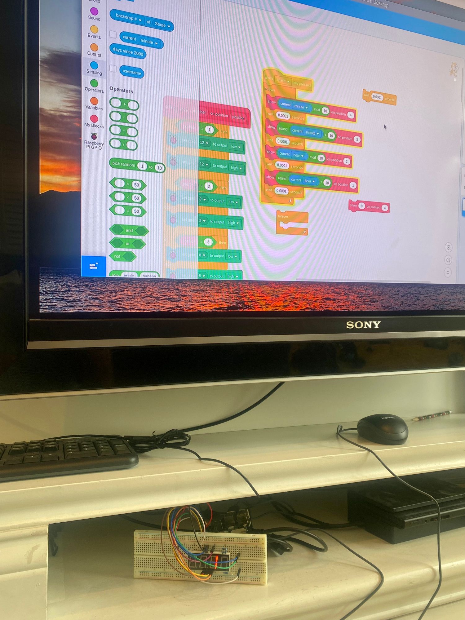 Wider shot of the breadboard showing that it's in a little cabinet under my TV, a mouse and keyboard above it. On the TV is a linux desktop running Scratch, and there's a fairly lengthy sequence of if-then's that sets the GPIO pins according to what digit the user is trying to display in what position.