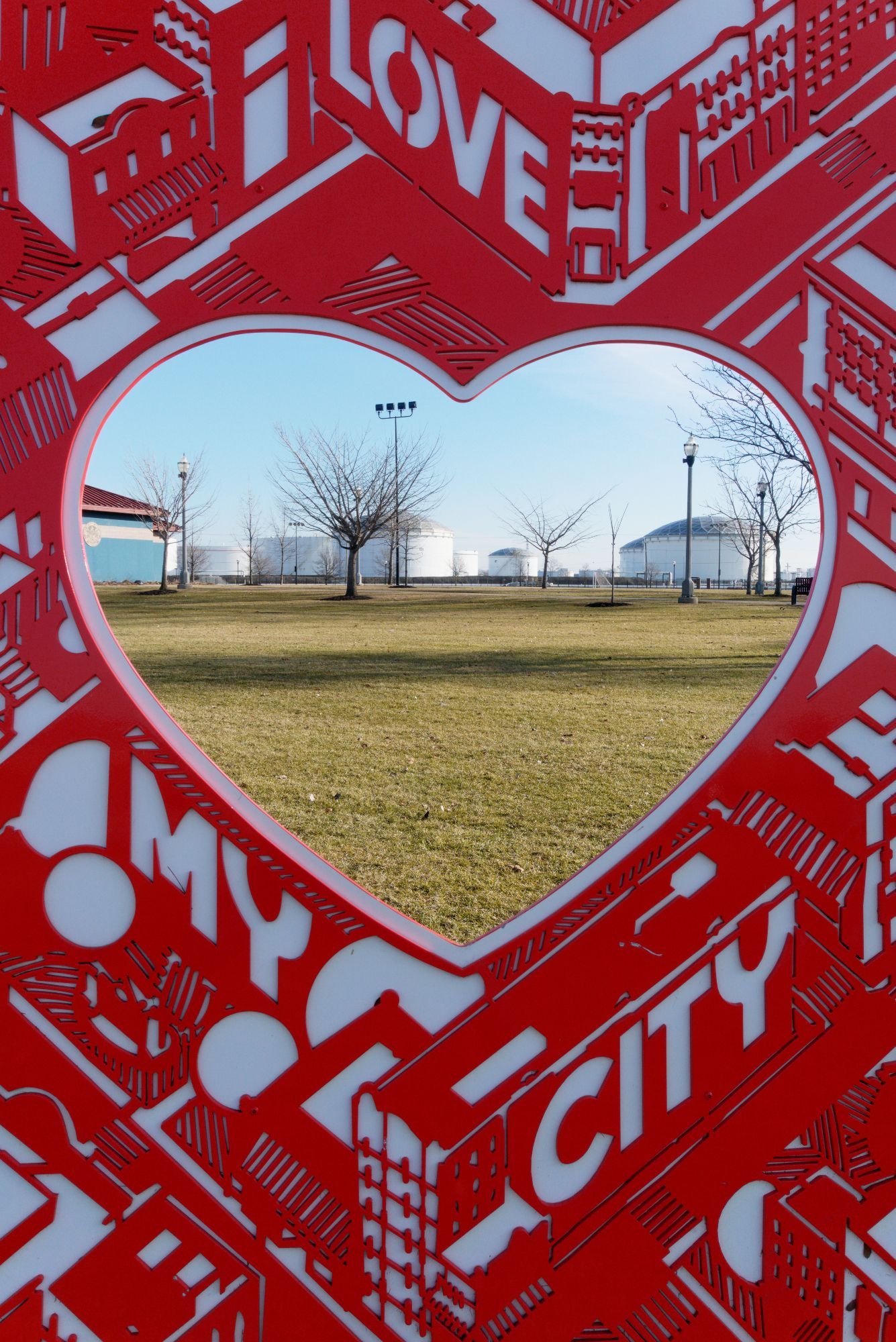 In the foreground is an art installation featuring the slogan "love my city". It's white on cherry red. In the center is a heart-shaped hole, letting you peer through. Through the hole, we see a grassy field, and beyond it (not very far) are more of these same white cylindrical petrochemical storage buildings.