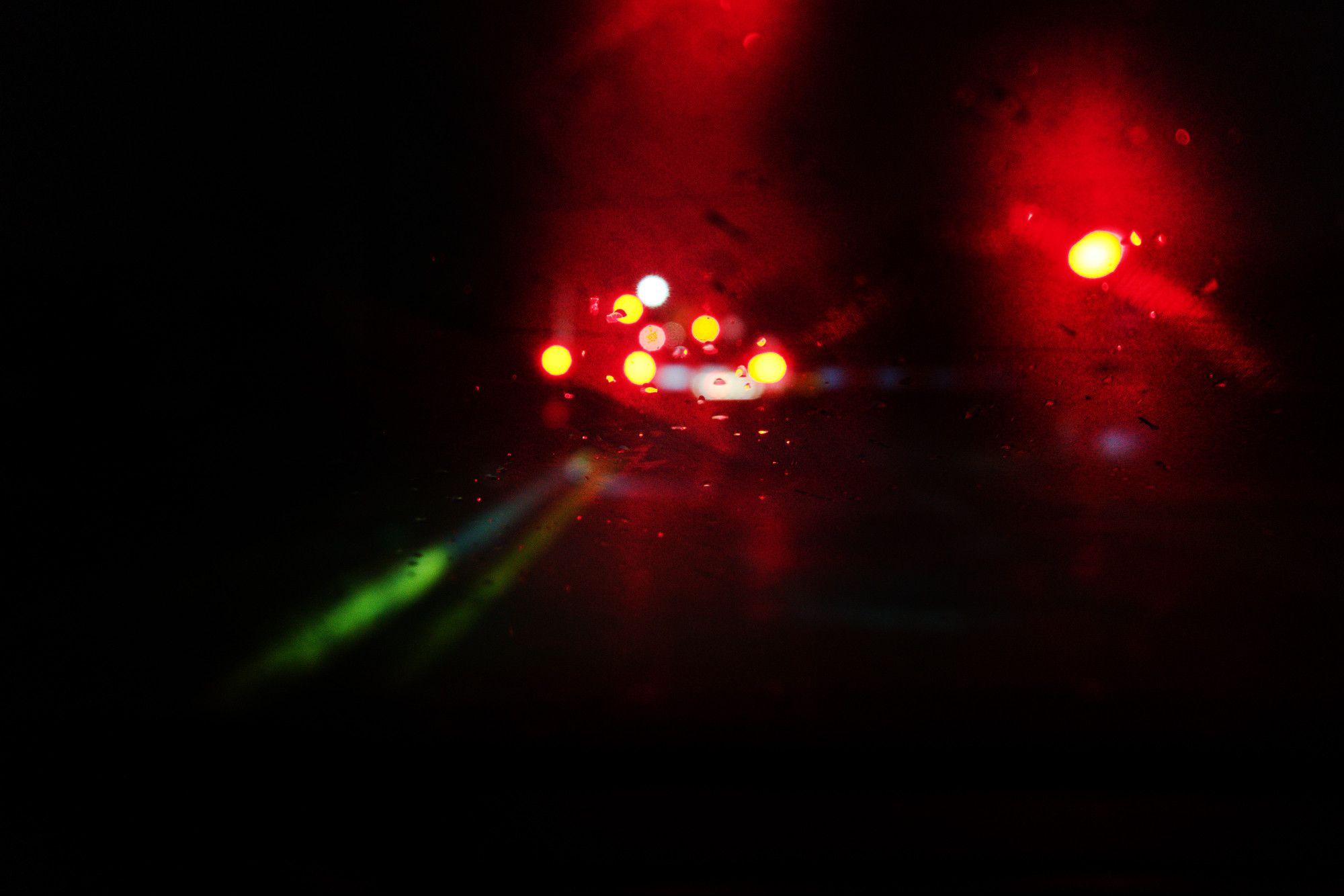 We're in a car. Outside it's pitch black and raining. We've stopped at a railroad crossing. The center yellow lines are semi-visible, going from bottom left of frame toward the center/horizon. Some red lights are bokeh'ed and bleeding because of the rain on the windshield.