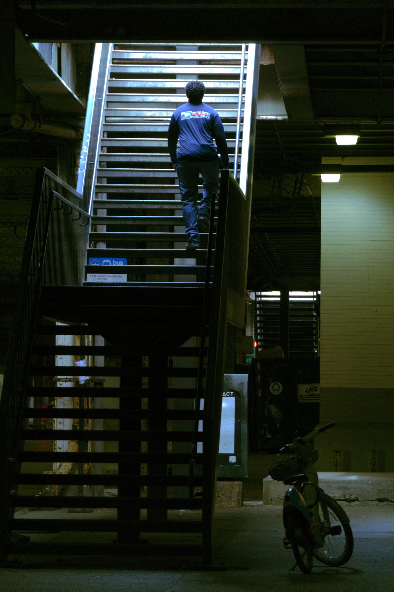 Here we're looking at a very similar, scene. But I think we're across the street on the east side, again looking north. This time a postal worker in blue attire is ascending the stairs. The light is just showing down on him, illuminating him from the top, while most of the rest of the scene is in shadows. On the right, some brick walls are illuminated in a greenish light.