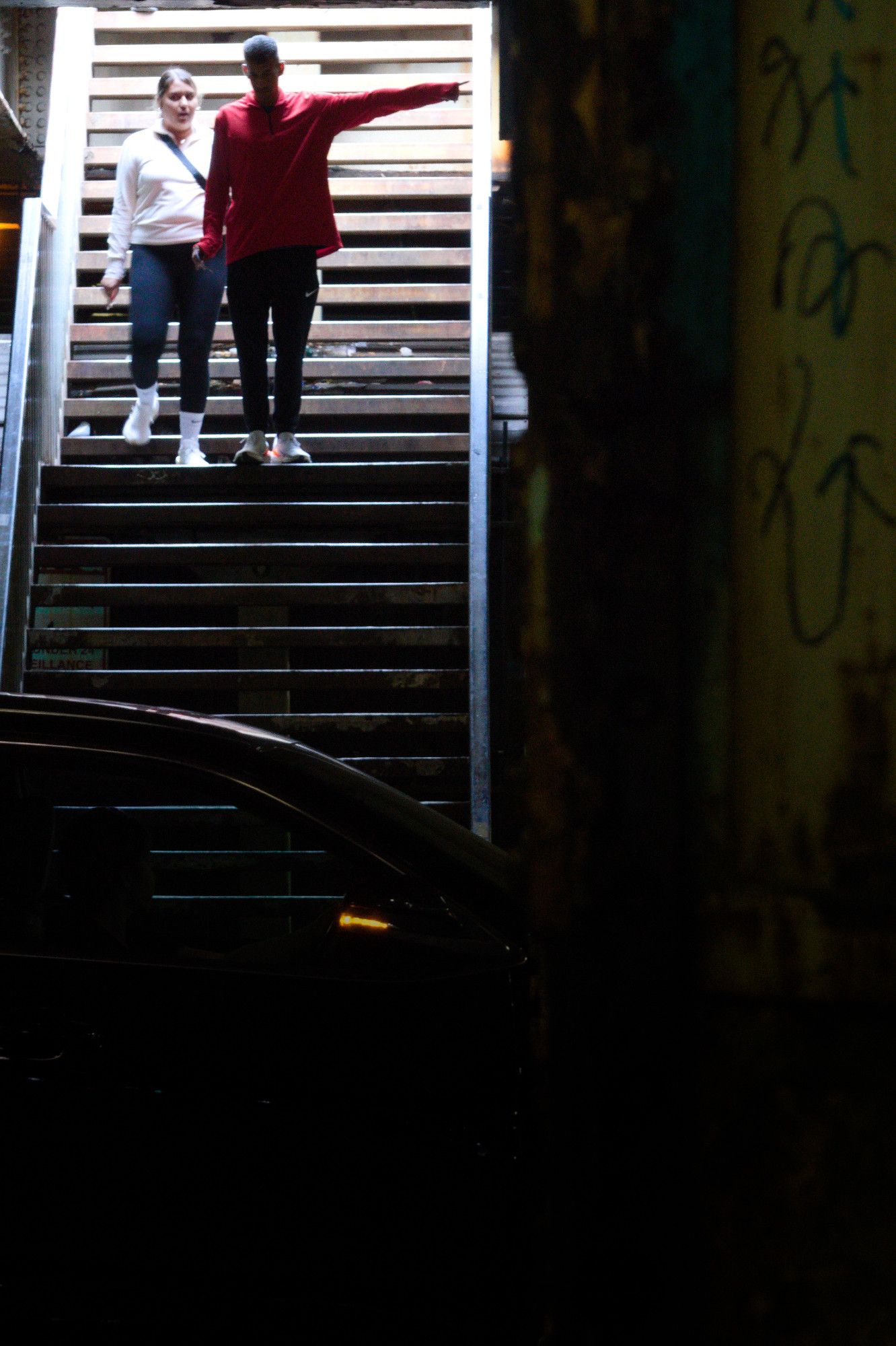 Same staircase, on the west side of the road, with two pedestrians descending the staircase. A woman in a white top and jeans descends behind a man in a red shirt and jeans. His left arm is extended, pointing (our) right in the image. It's not clear if he's giving directions, pointing out something noteworthy, etc.