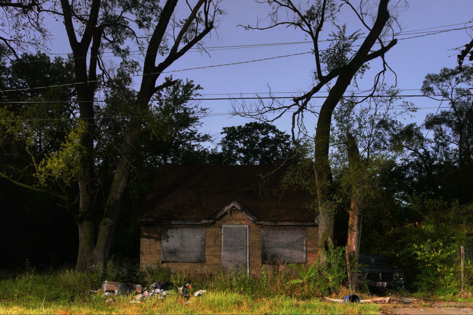 We're looking head-on at a probably-abandoned abandoned house. The house is framed by a pair of probably-dead trees on either side. To the right of the house, a vehicle is parked in the shadows at the back of the driveway. Powerlines streak left-to-right above the house. The sidewalk and street are cropped out of this picture, giving the house the appearance of standing alone in a field, rather than at the end of a dead-end residential street.