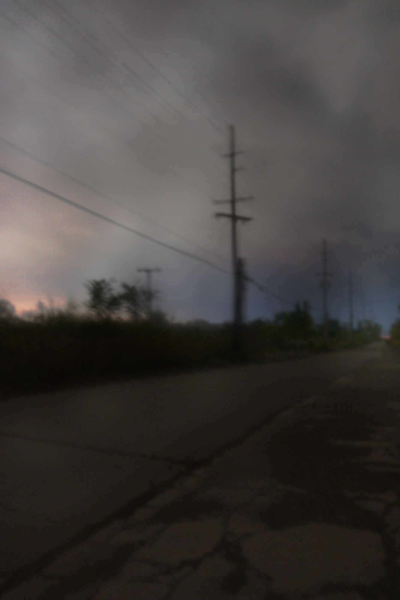 We're on a narrow road in the woods. On our left, a string of power lines cascade from the left side of the image, off into the distance, to the right side. Beyond it, some scraggly trees appear as silhouettes against the nigh sky. The night sky is creamy clouds with reflected city lights from beyond the forest. Some patches of peach colors warm the clouds on the bottom and left. We're looking through a wet window, which gives the image a dream-like, hazy quality that feels like a fuzzy, subjective memory rather than a crisp, objective recording of a moment.