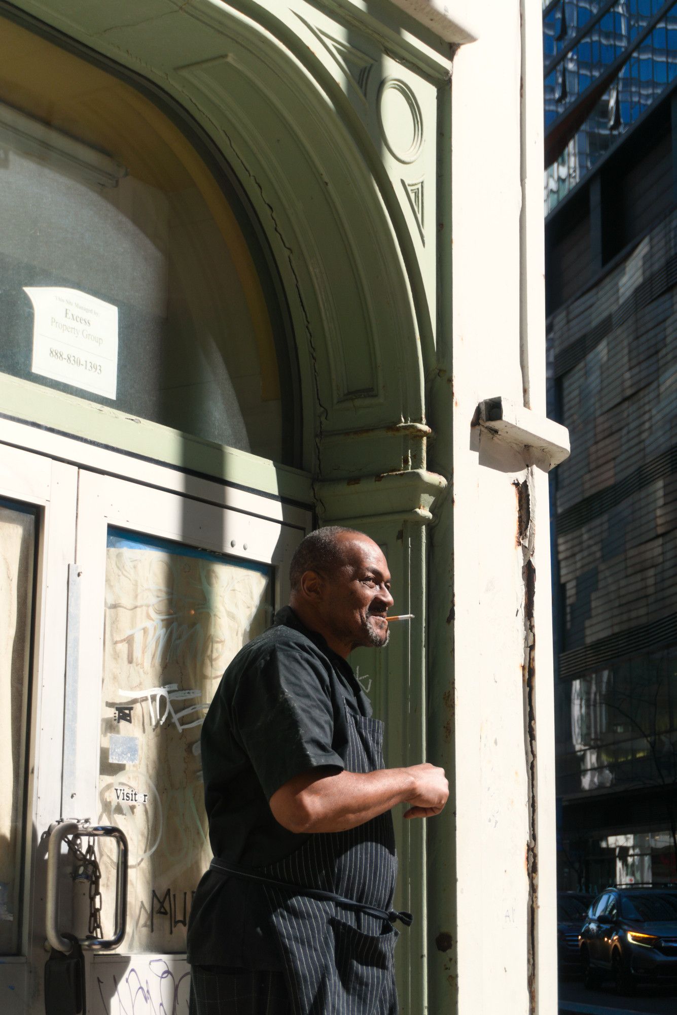 It's a sunny day in March. It's just after noon, and the harsh midday light is directly on this scene on the NE corner of an intersection in the loop. A man in a black shirt and a black pinstripe apron stands outside a building during a a smoke break. The corner of the building he stands in has an arch starting over his right and exiting up over him on the left of the frame.