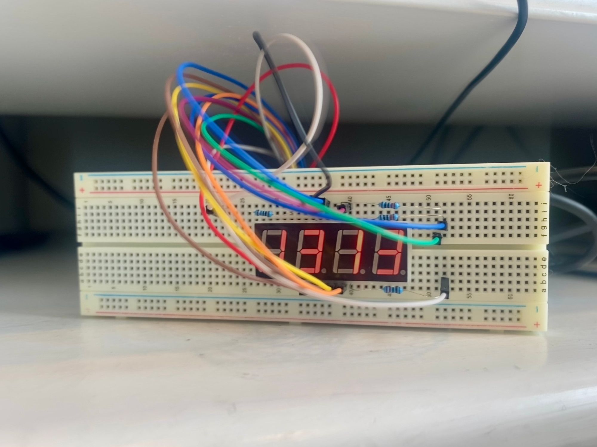 A breadboard is propped up on its side, showing a 4-digit, 7-segment display that reads "13:12" (military time for an hour and 12 minutes past noon). The board has 4 blue resistors connecting things, and about a dozen jumper wires leading back to the raspberry pi, which is behind the breadboard.