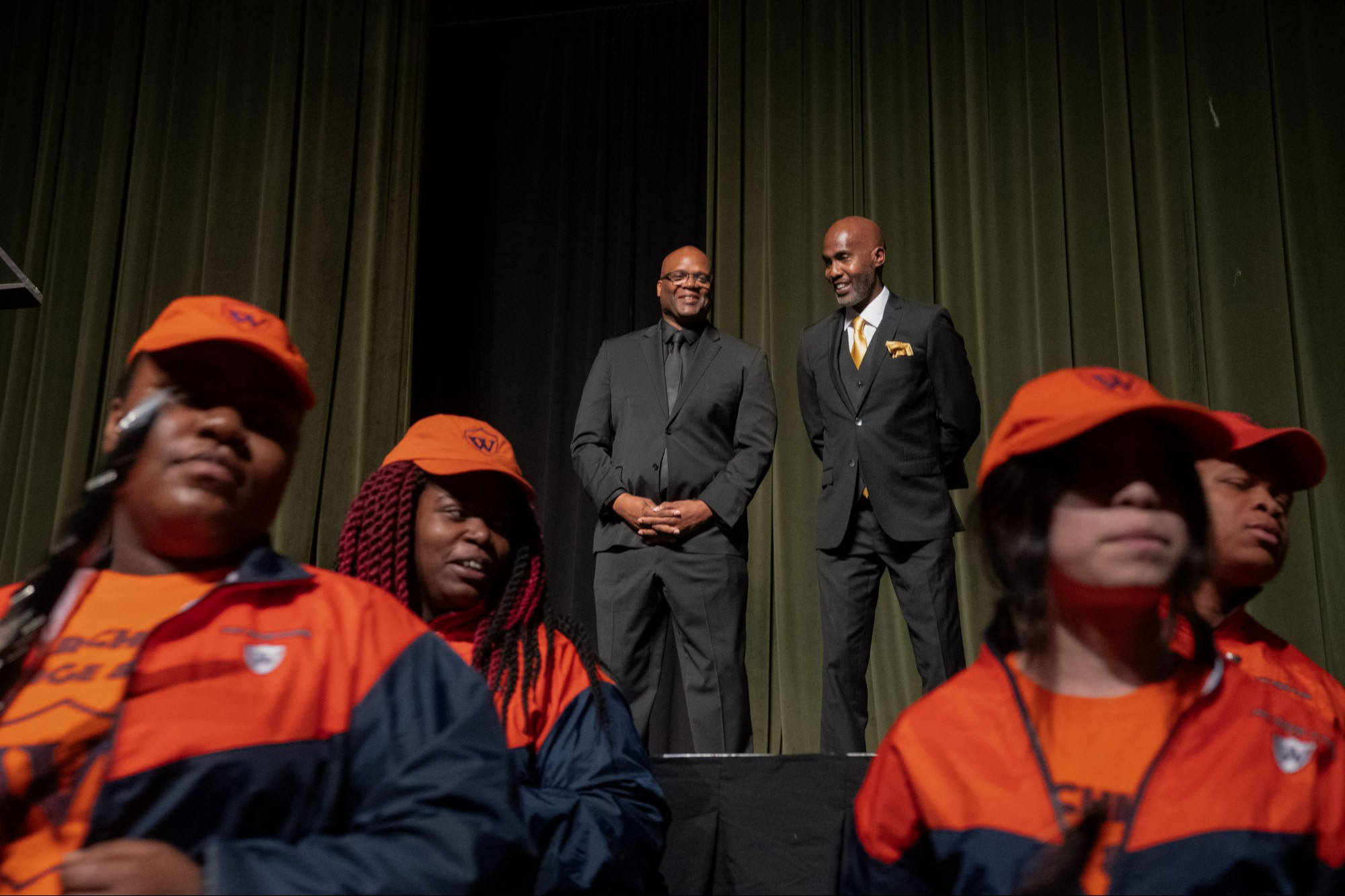 two people stand on stage in the background with marching band high school students stand close in the foreground