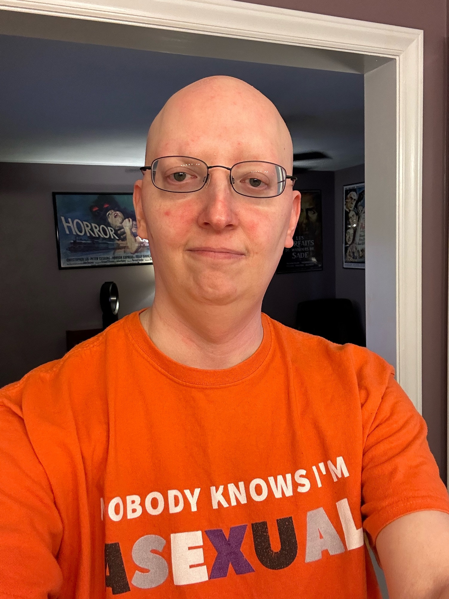 Selfie of me wearing glasses and my Ace Pride T-shirt for Ace week that says “Nobody Knows I’m Asexual”. With my movie poster for Horror Express in the background.